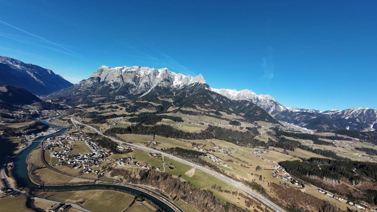 Panoramische Luftaufnahme eines weiten Tals mit schneebedeckten Bergen, einer Stadt und einer Autobahn.