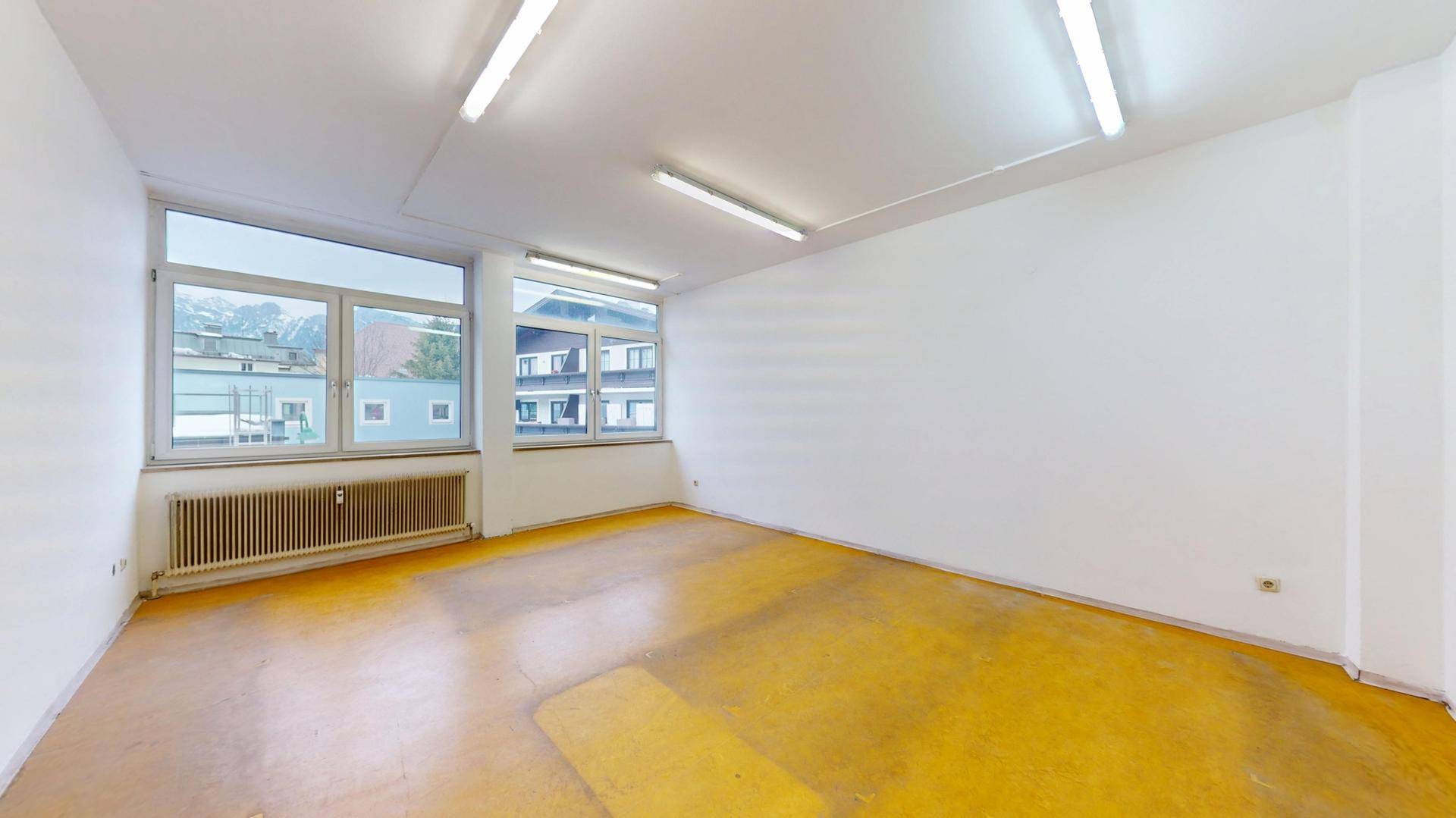 Geräumiger Büroraum mit gelbem Boden, weißen Wänden und Fenstern mit Blick auf die Umgebung.