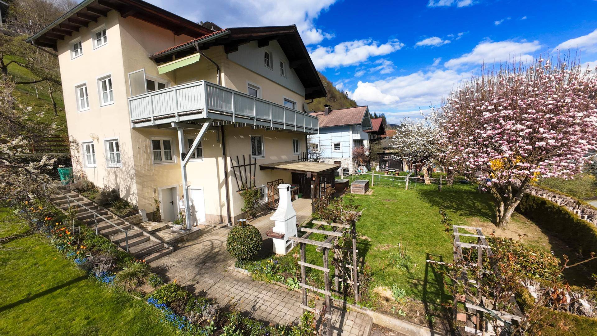 Hausansicht von der Gartenseite mit blühenden Bäumen und gepflegter Rasenfläche.
