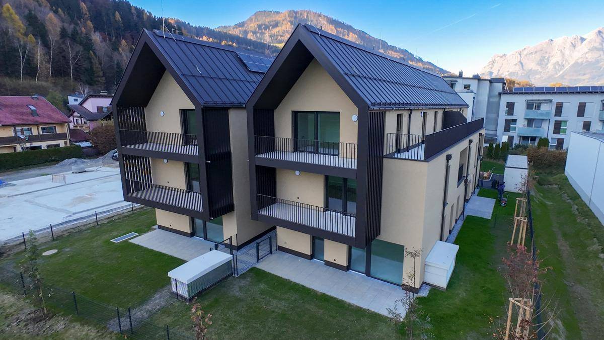 Modernes Mehrfamilienhaus mit Satteldach und Balkonen, umgeben von grüner Landschaft und Bergen.