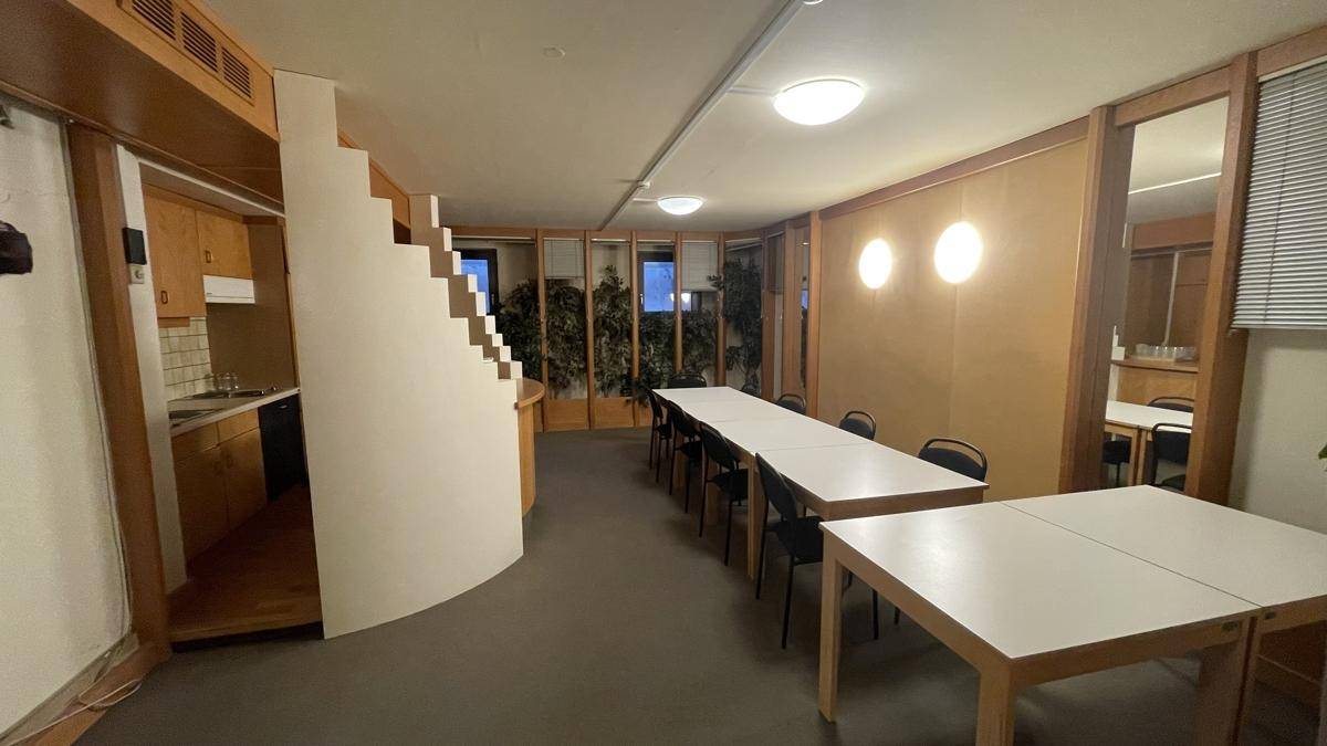 Pausenraum mit langer Tischreihe, Stühlen und einer kleinen Küchenzeile, ideal für Mitarbeiter.