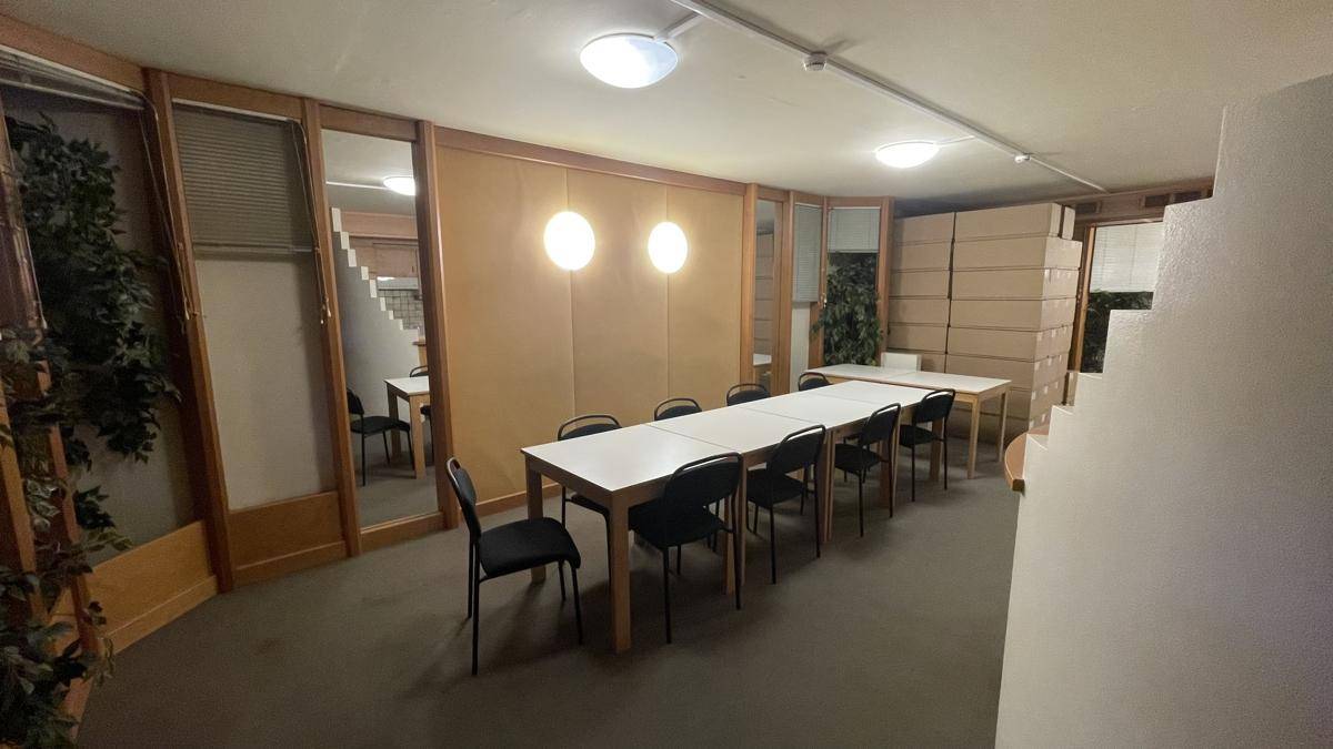 Pausenraum mit langen Tischen und Stühlen, umgeben von Holzverkleidung und Pflanzen, bietet Platz für Erholung.