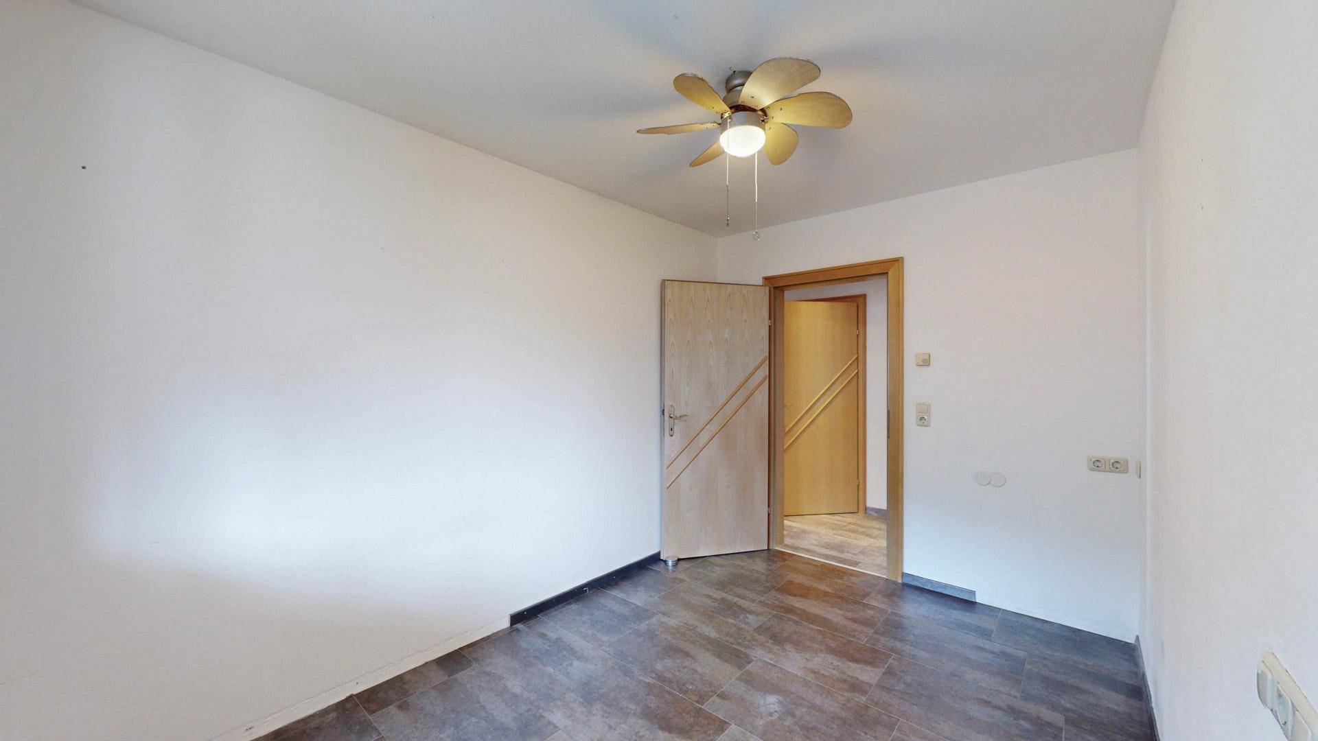 Leeres Zimmer mit dunklem Fliesenboden und Deckenventilator, bereit zur individuellen Gestaltung.