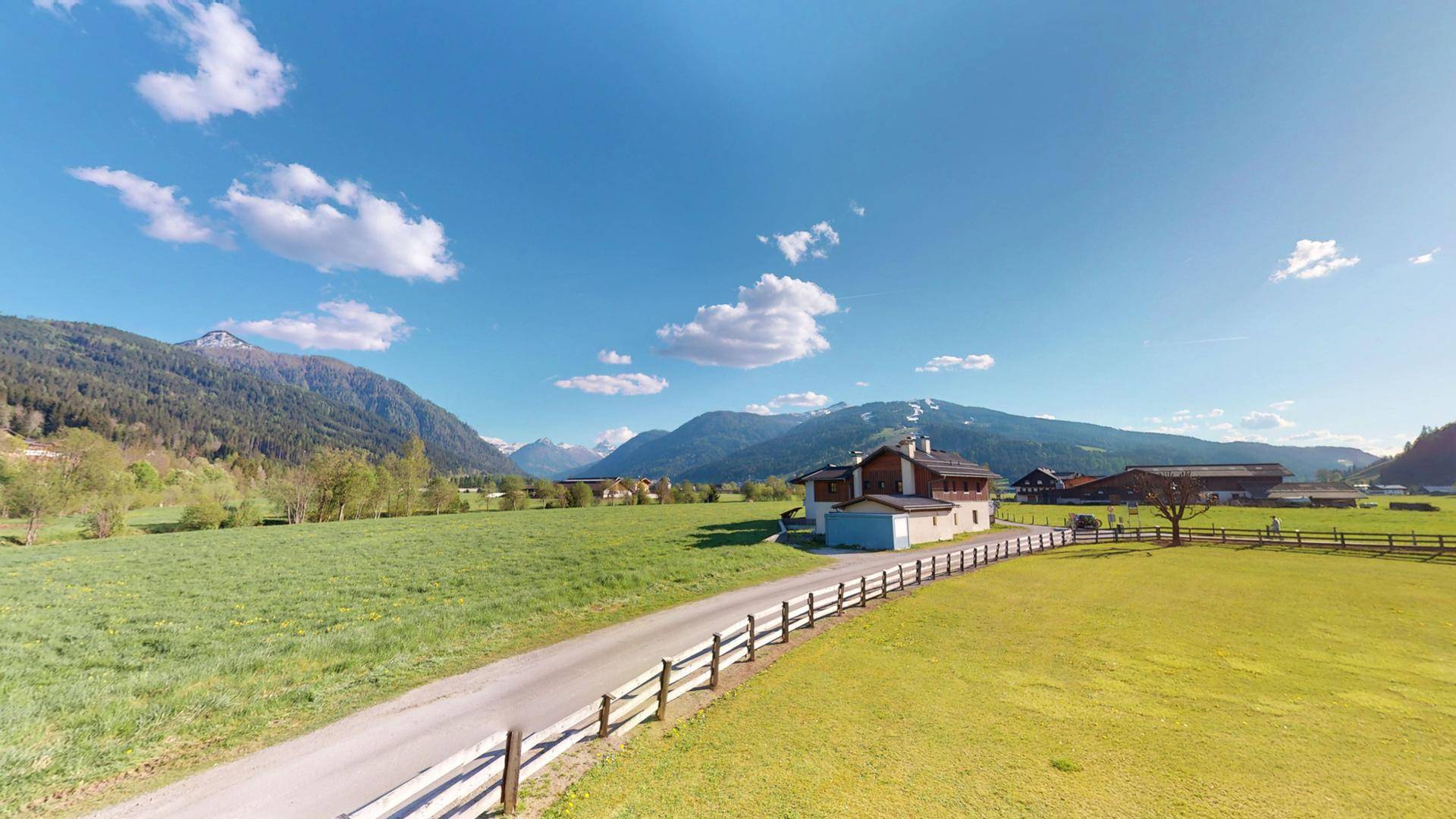 Weitläufige Landschaft mit grünen Wiesen, Bergen und einem traditionellen Gebäude unter blauem Himmel.