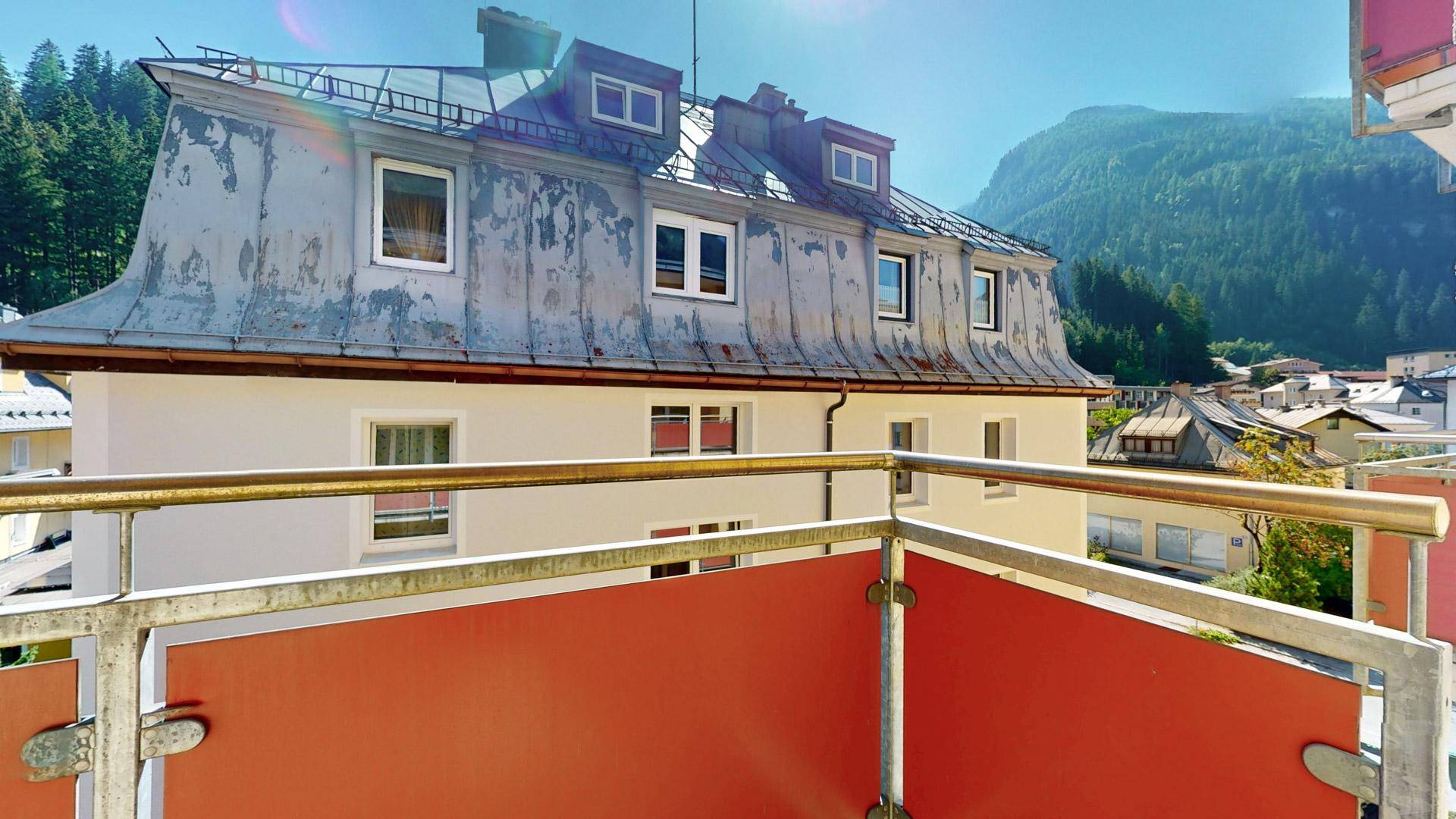 Balkon mit Aussicht auf ein charmantes Gebäude mit Mansarddach und die umliegende Berglandschaft.