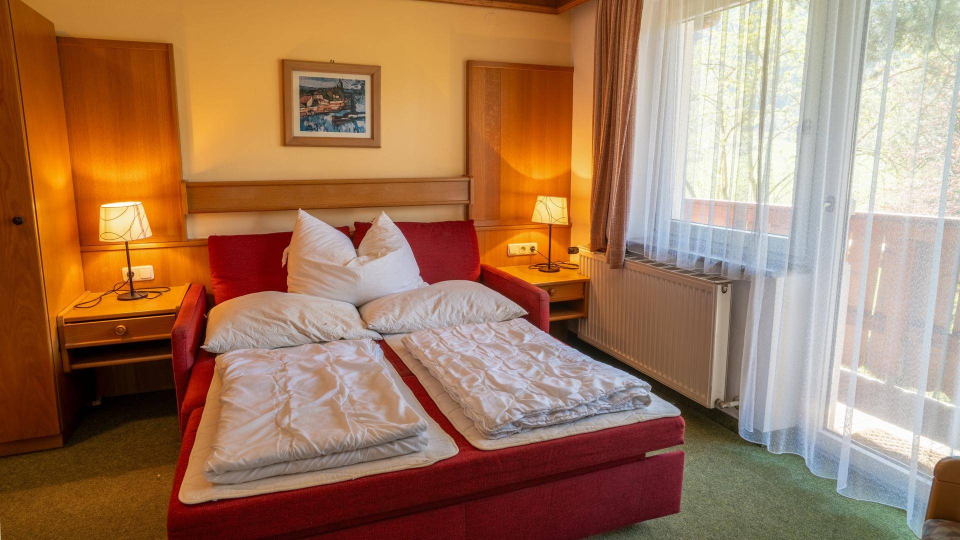Helles Schlafzimmer mit rotem Schlafsofa, Holzmöbeln und Zugang zum Balkon.