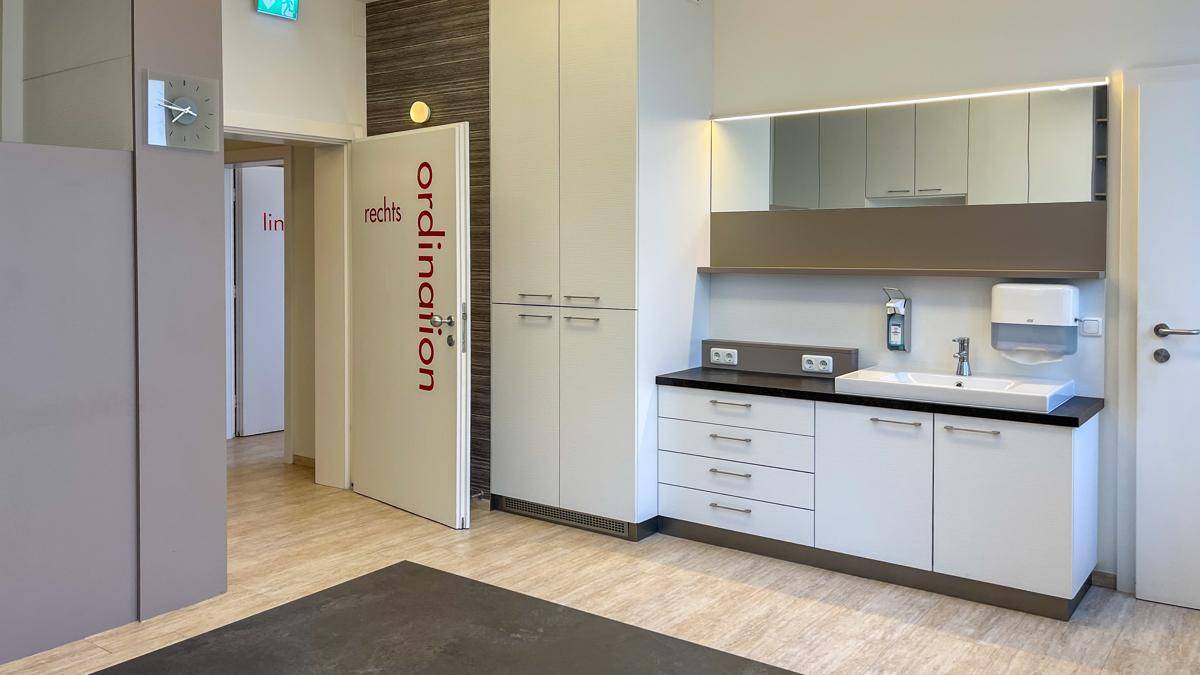Heller Flur mit einem Waschbeckenbereich, Spiegelschrank und Zugang zu weiteren Praxisräumen.