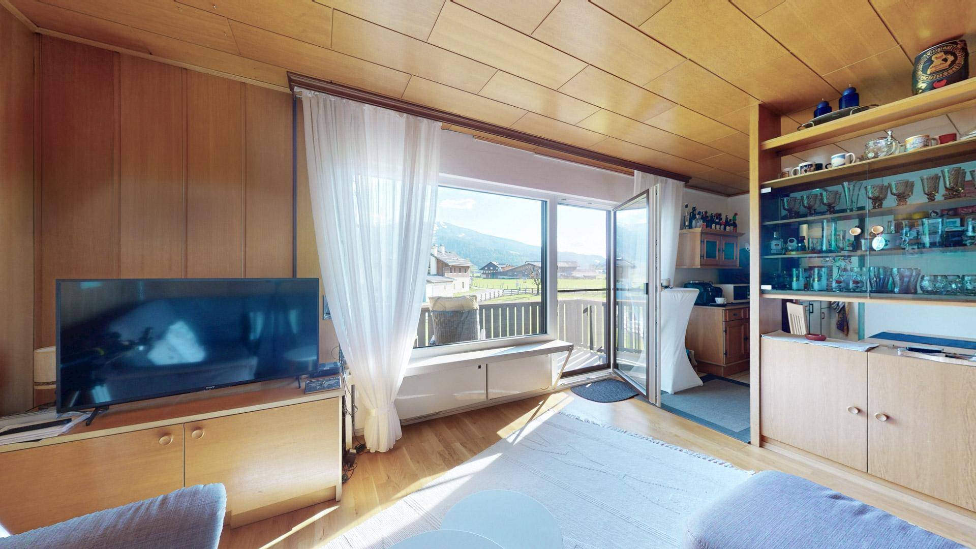 Helles Wohnzimmer mit Zugang zum Balkon, Holzvertäfelung und Blick auf die umliegende Natur.