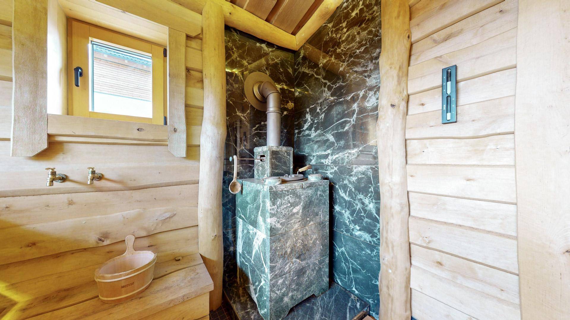Detailansicht des Saunaofens aus dunklem Marmor mit traditionellem Aufgusszubehör und Holzwänden.