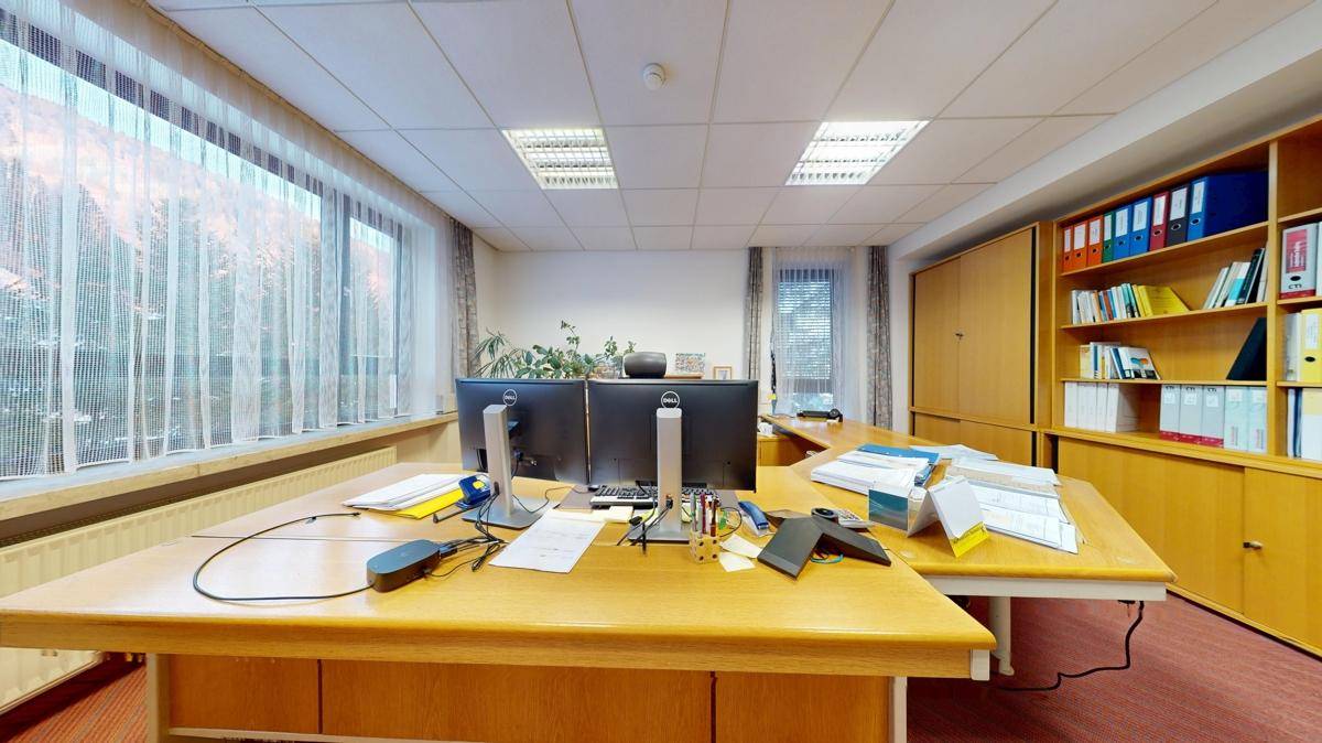 Büro mit Teppichboden, zwei Monitoren auf dem Schreibtisch und Holzregalen, bereit für den Arbeitsalltag.