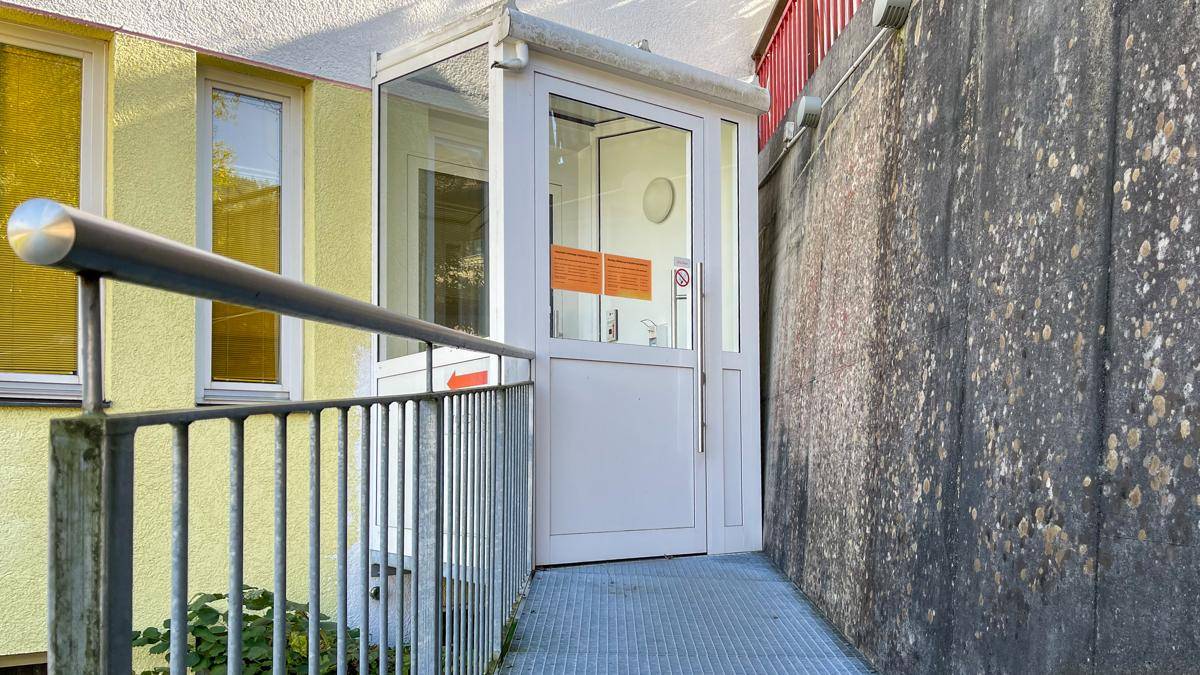 Eingangsbereich mit Metallrampe und Handlauf für barrierefreien Zugang zum Gebäude, umgeben von Grün.