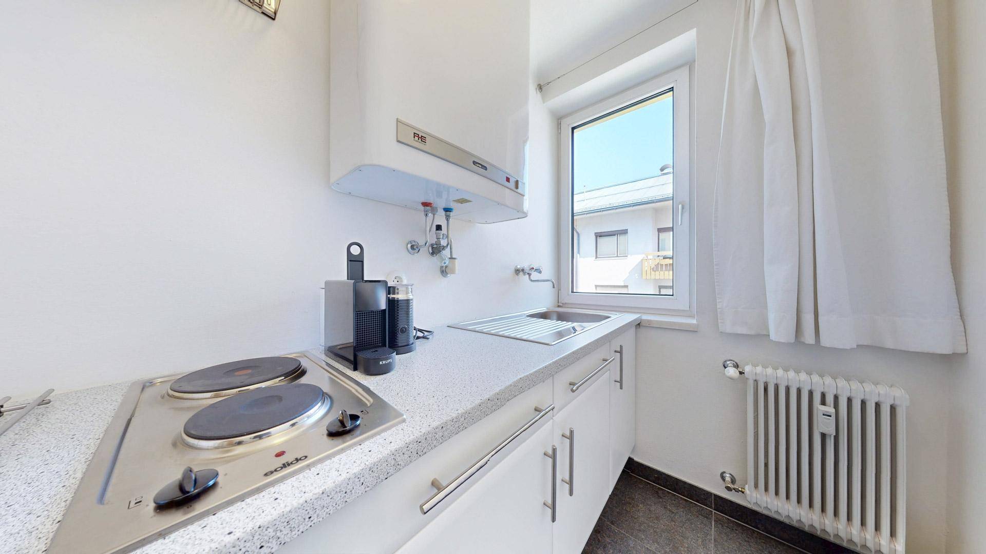 Funktionale Küche mit weißen Unterschränken, Elektrokochfeld und Spüle unter dem Fenster.