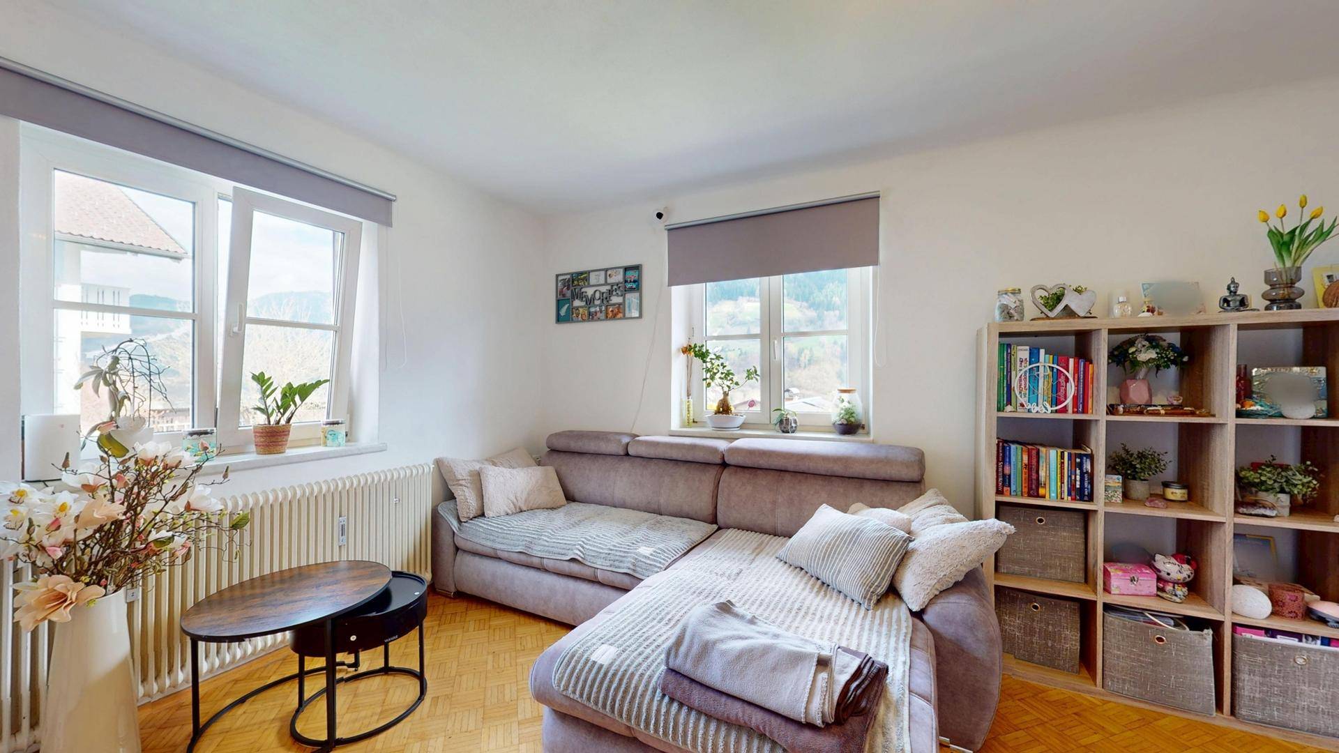 Helles Wohnzimmer mit Ecksofa, Bücherregal und Fenstern mit Blick ins Grüne.