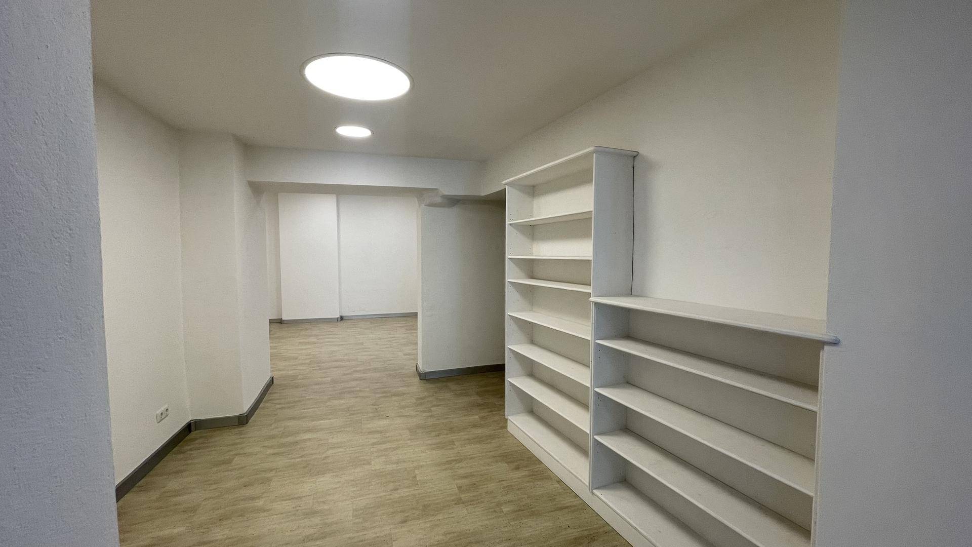 Innenraum mit Holzoptikboden, weißen Wänden, Einbauregalen und runden Deckenleuchten.