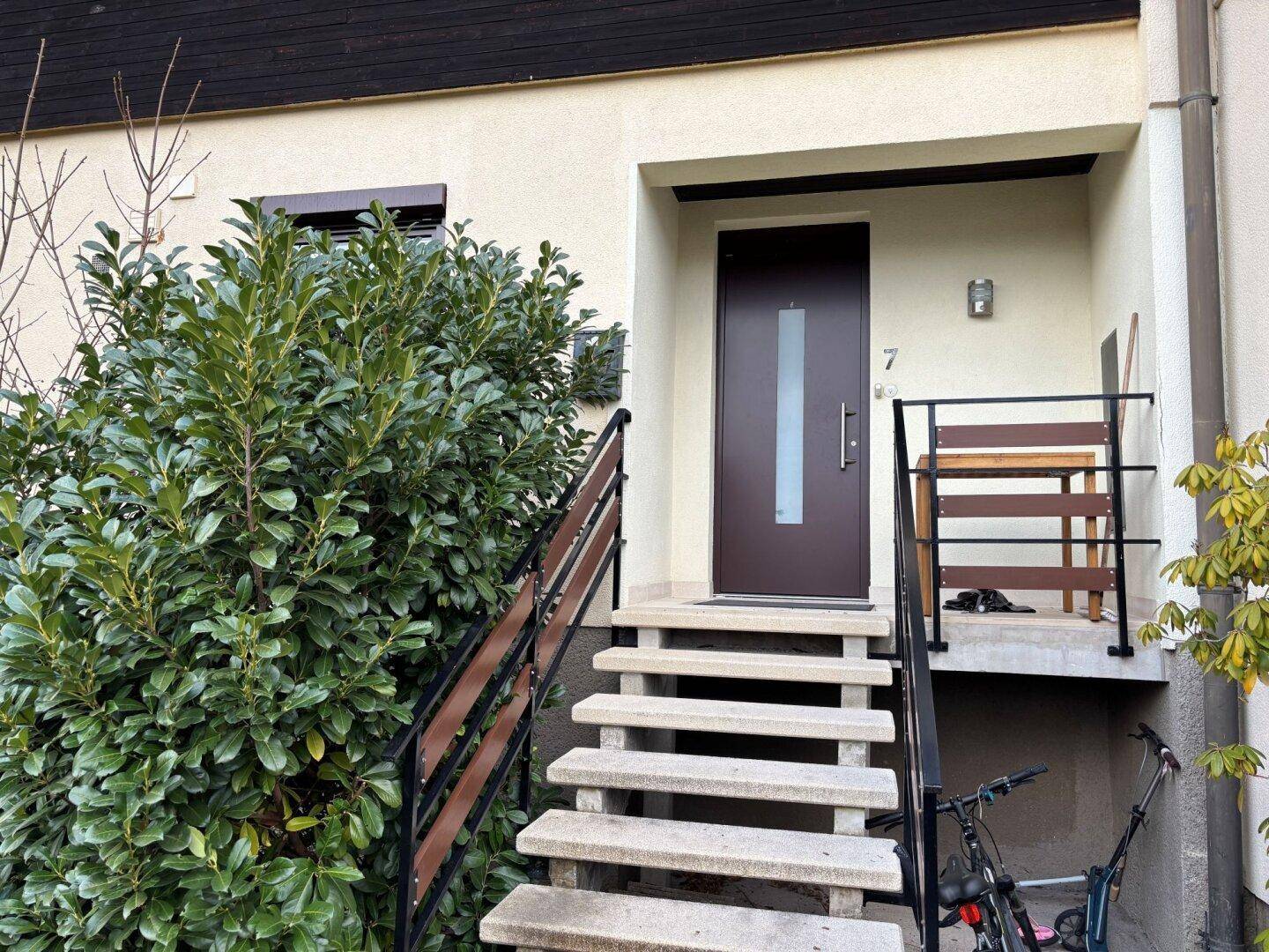 Eingangsbereich eines Reihenhauses mit moderner Haustür, Steintreppe und grüner Bepflanzung.