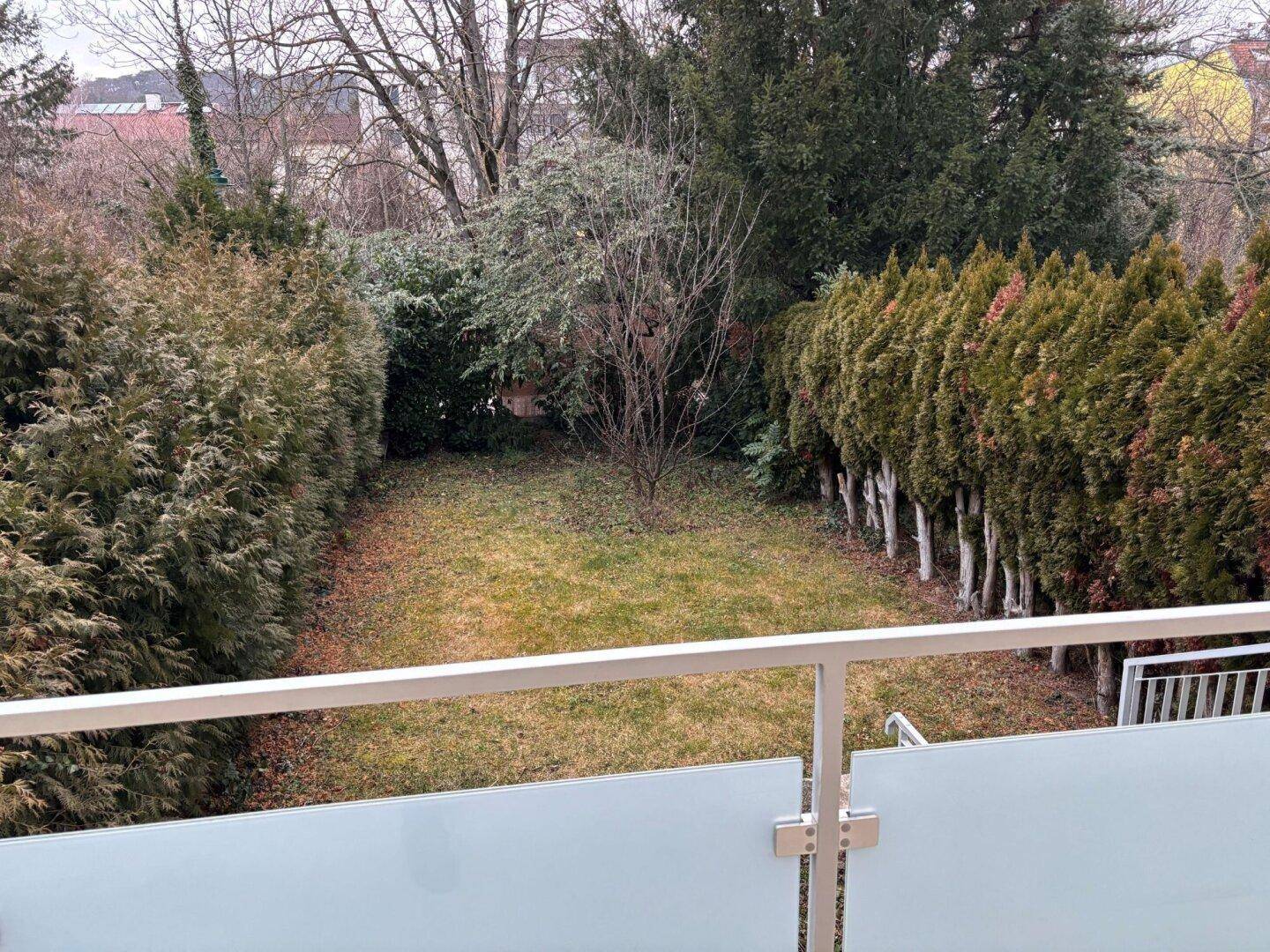 Blick vom Balkon auf den privaten Gartenbereich mit Rasenfläche und hohen Hecken.