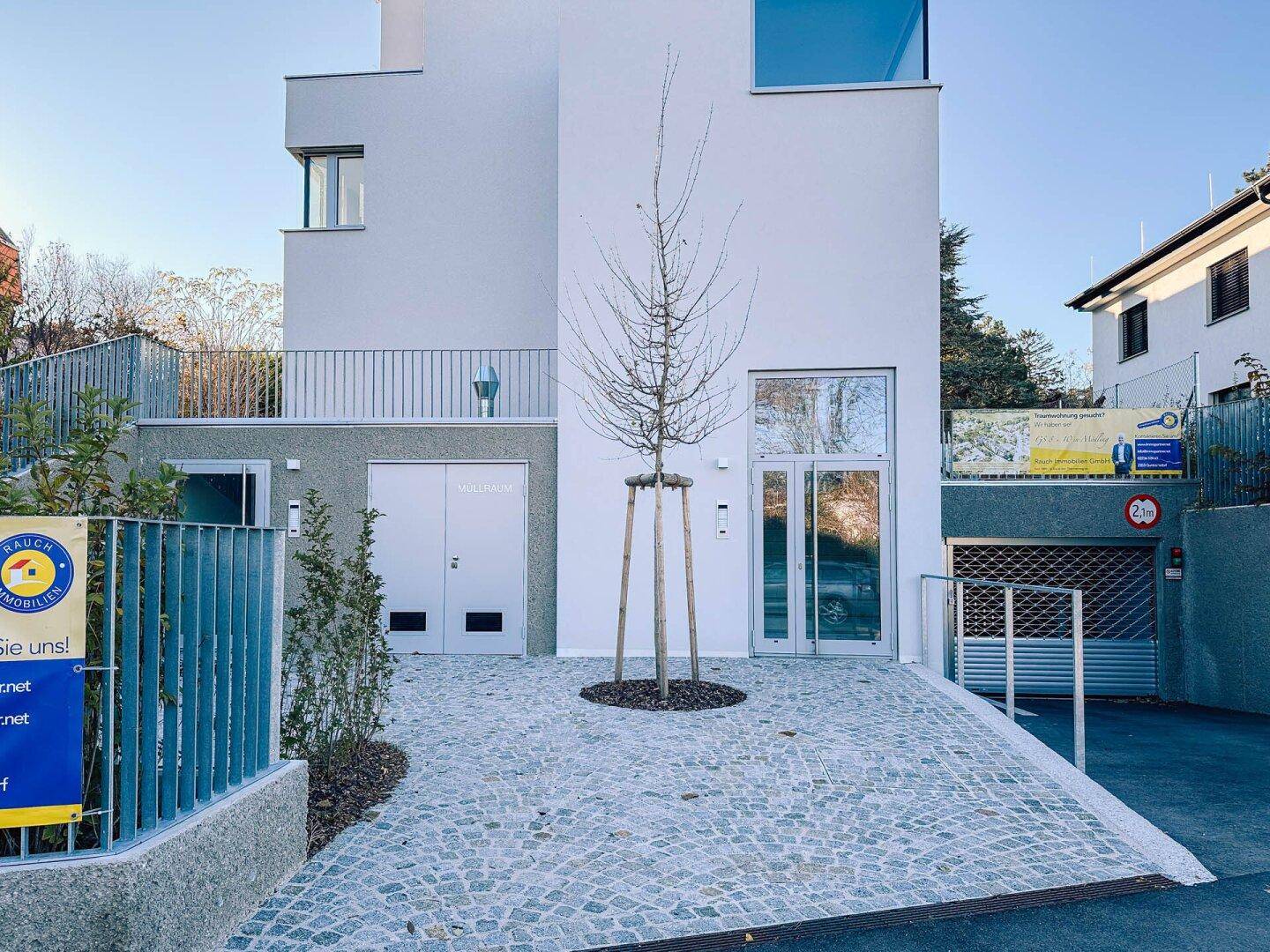 Eingangsbereich des modernen Gebäudes mit gepflastertem Vorplatz und Zufahrt zur Tiefgarage.