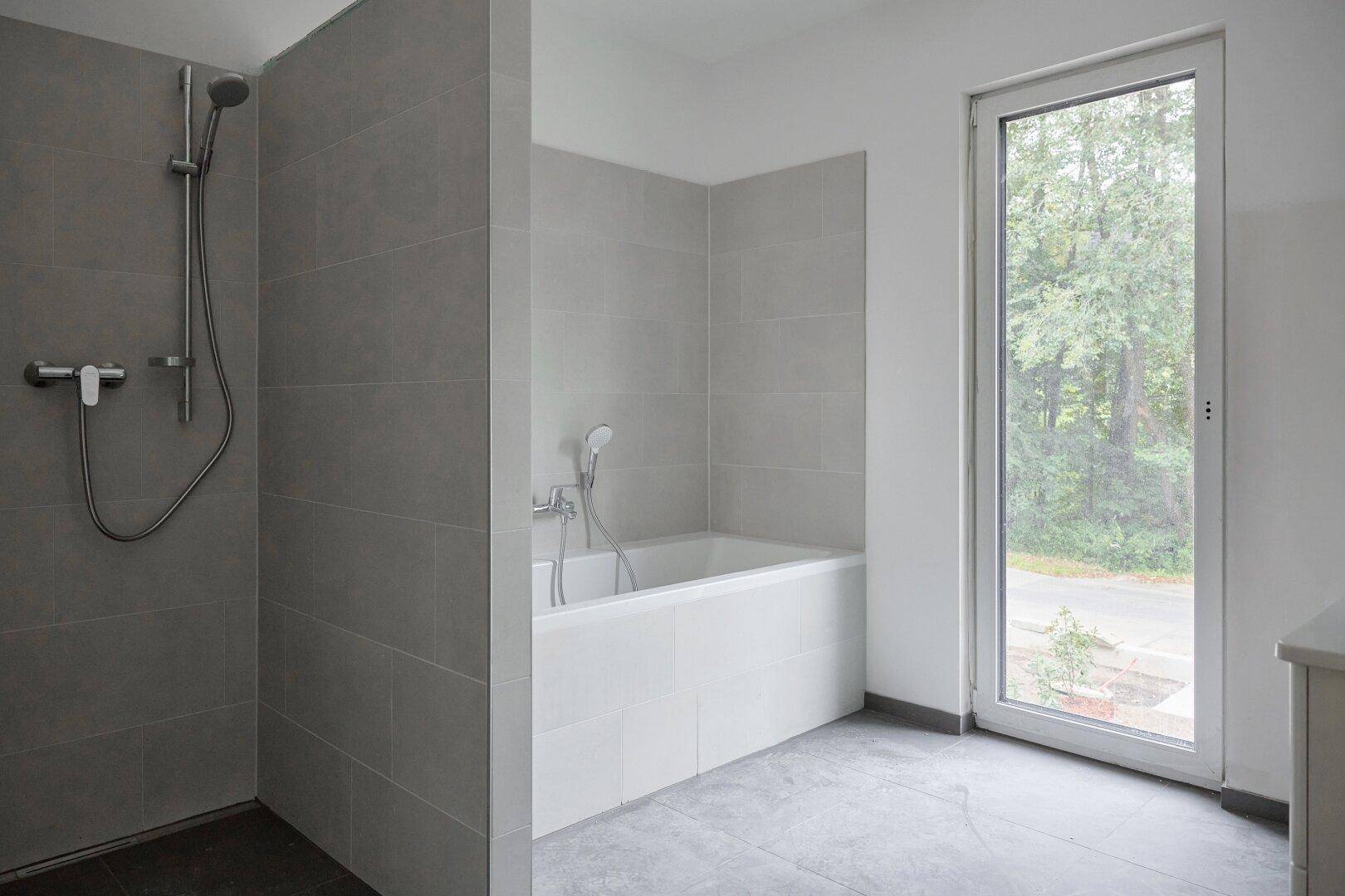 Helles Badezimmer mit Badewanne, Duschbereich und großer Fenstertür zum Außenbereich.