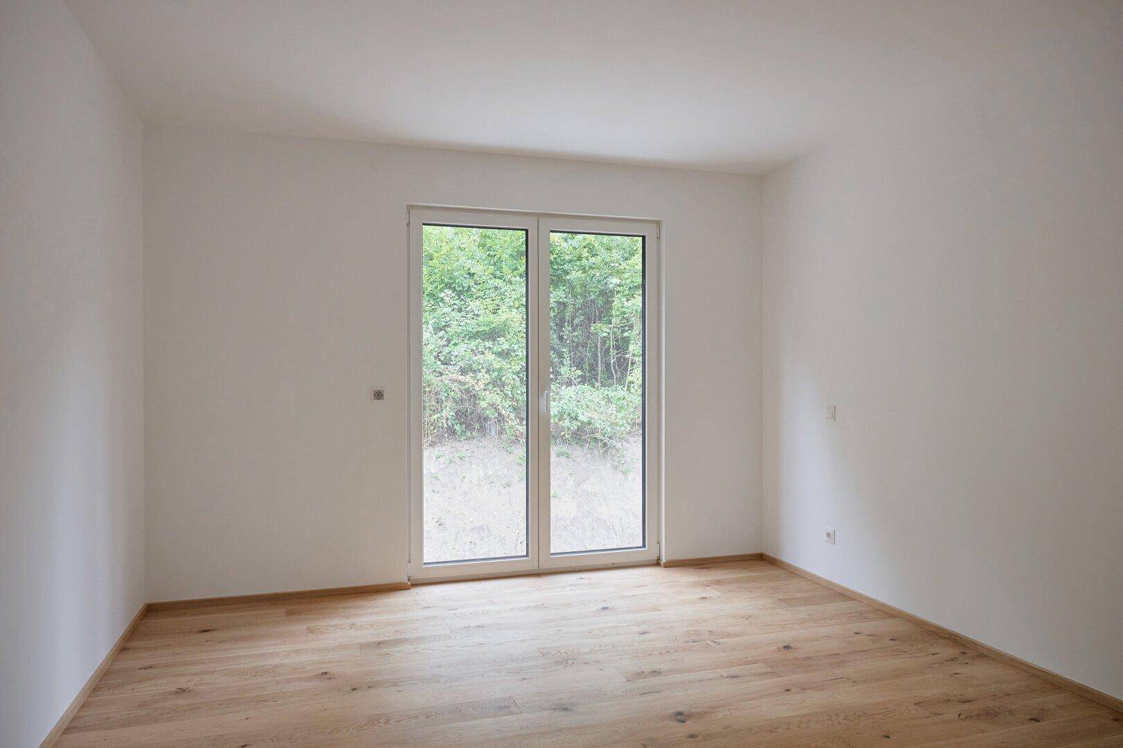 Leeres Zimmer mit hellem Holzboden und großer Fenstertür, die viel Tageslicht bietet.