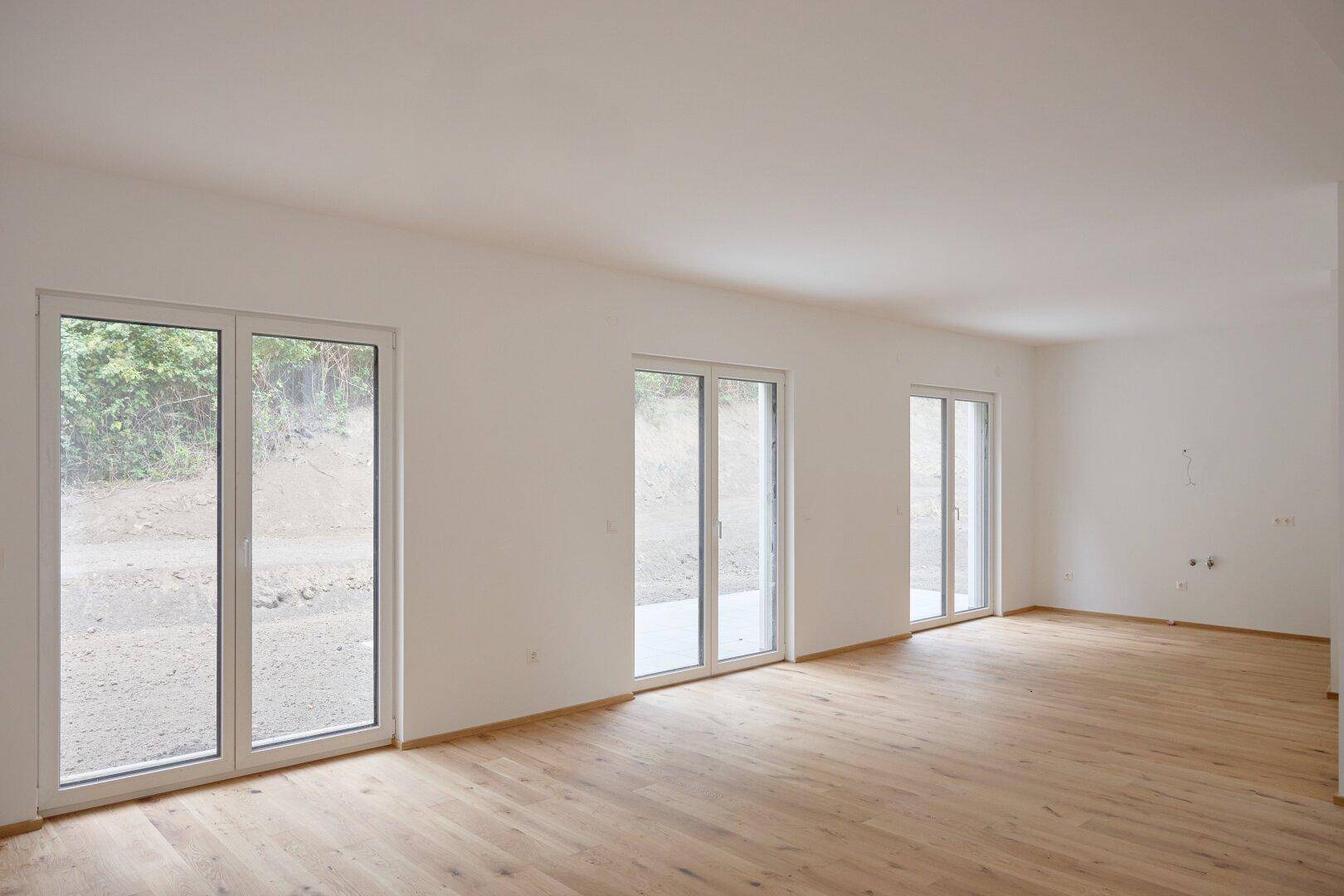 Unmöbliertes Wohnzimmer mit hellem Holzboden und mehreren Fenstertüren zum Außenbereich.