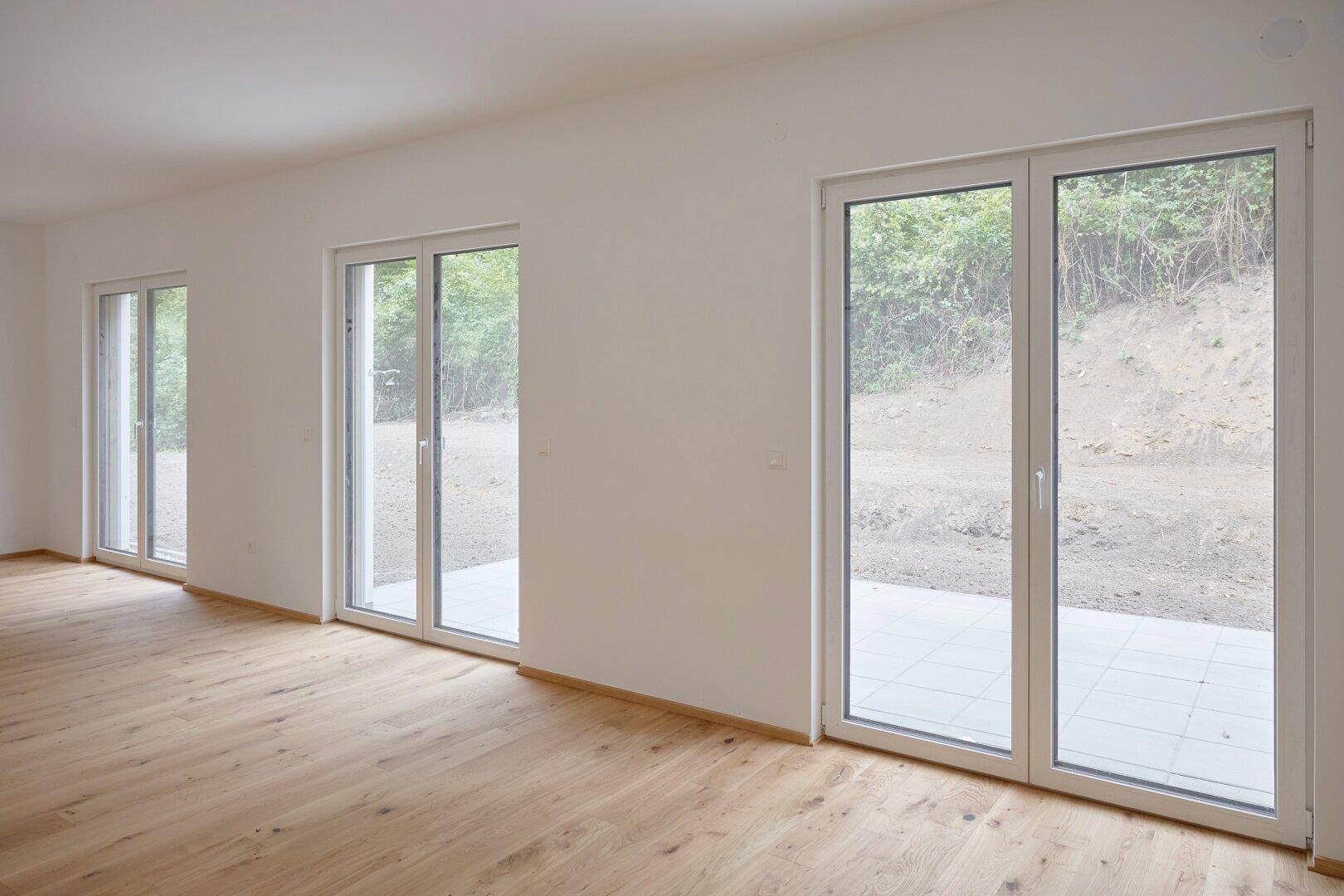 Geräumiges Wohnzimmer mit hellem Holzboden und drei großen Fenstertüren zum Garten.