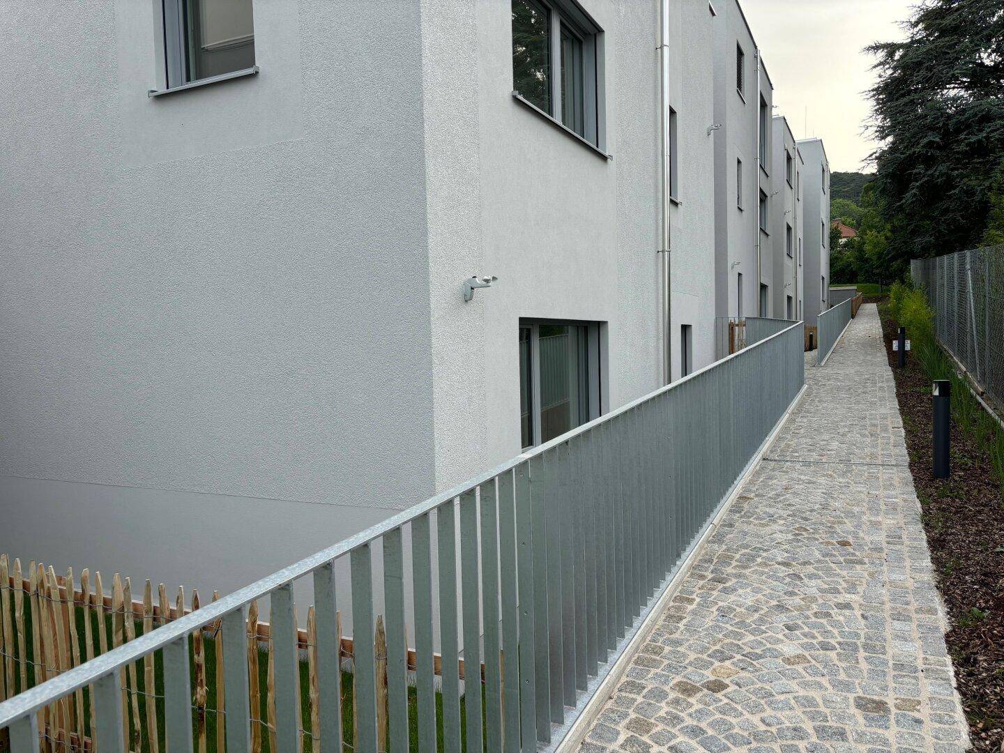 Gepflasterter Gehweg entlang der modernen Wohngebäude, umgeben von einer Holzumzäunung.