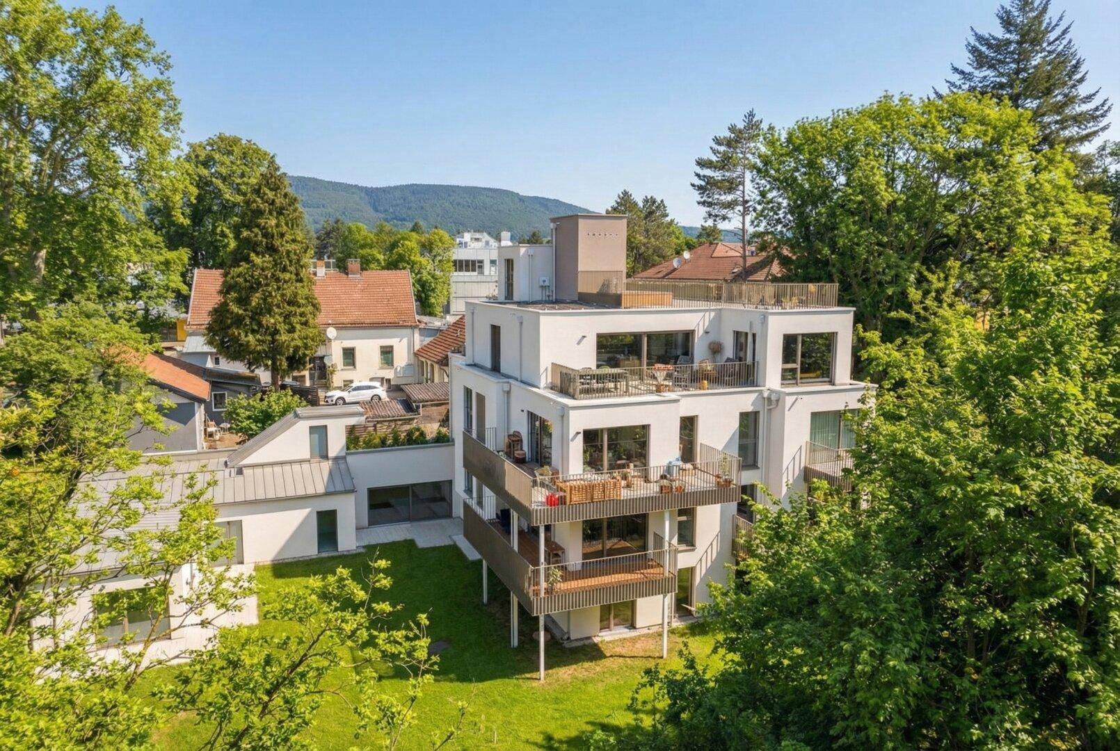 Modernes Mehrfamilienhaus mit mehreren Balkonen und Dachterrasse, umgeben von viel Grün.