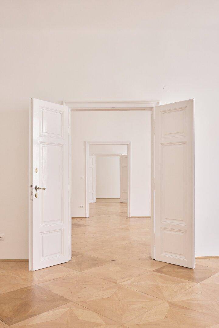 Blick durch eine Reihe offener Türen in helle, leere Räume mit klassischem Parkettboden und hohen Decken.