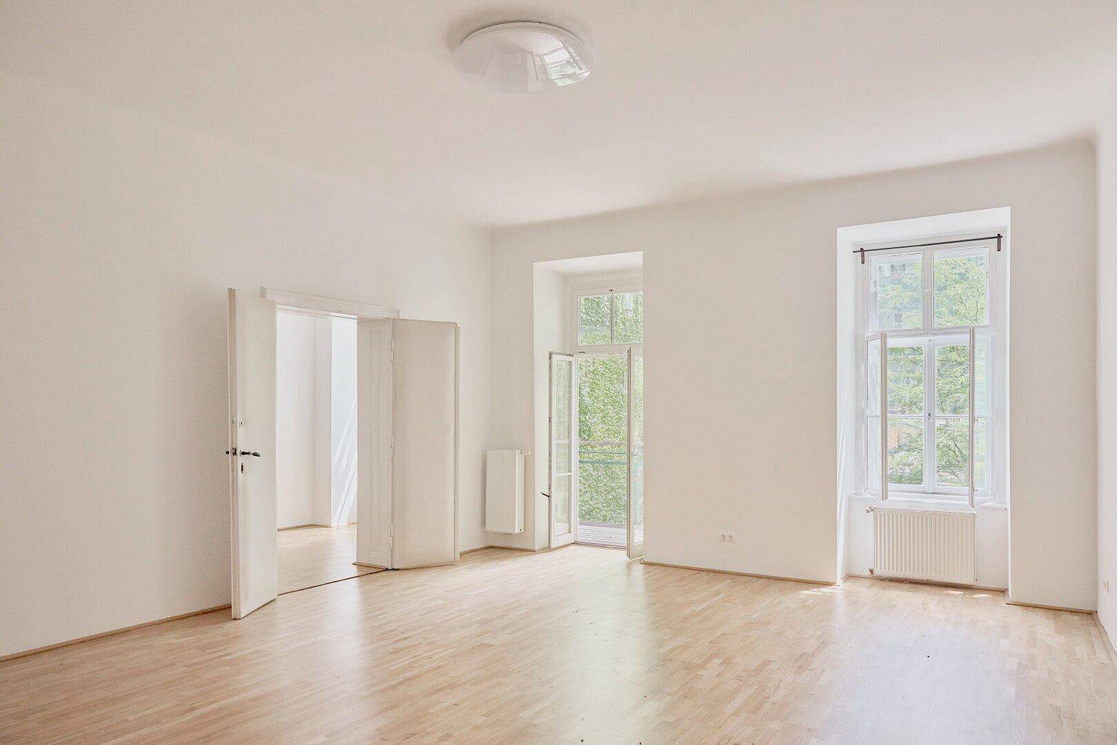 Heller, leerer Raum mit Holzdielenboden, zwei Fenstern und offenem Durchgang zu einem weiteren Zimmer.