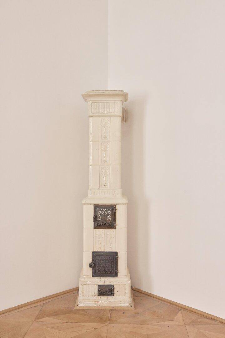 Ein traditioneller weißer Kachelofen steht in der Ecke eines hellen Zimmers mit Parkettboden.