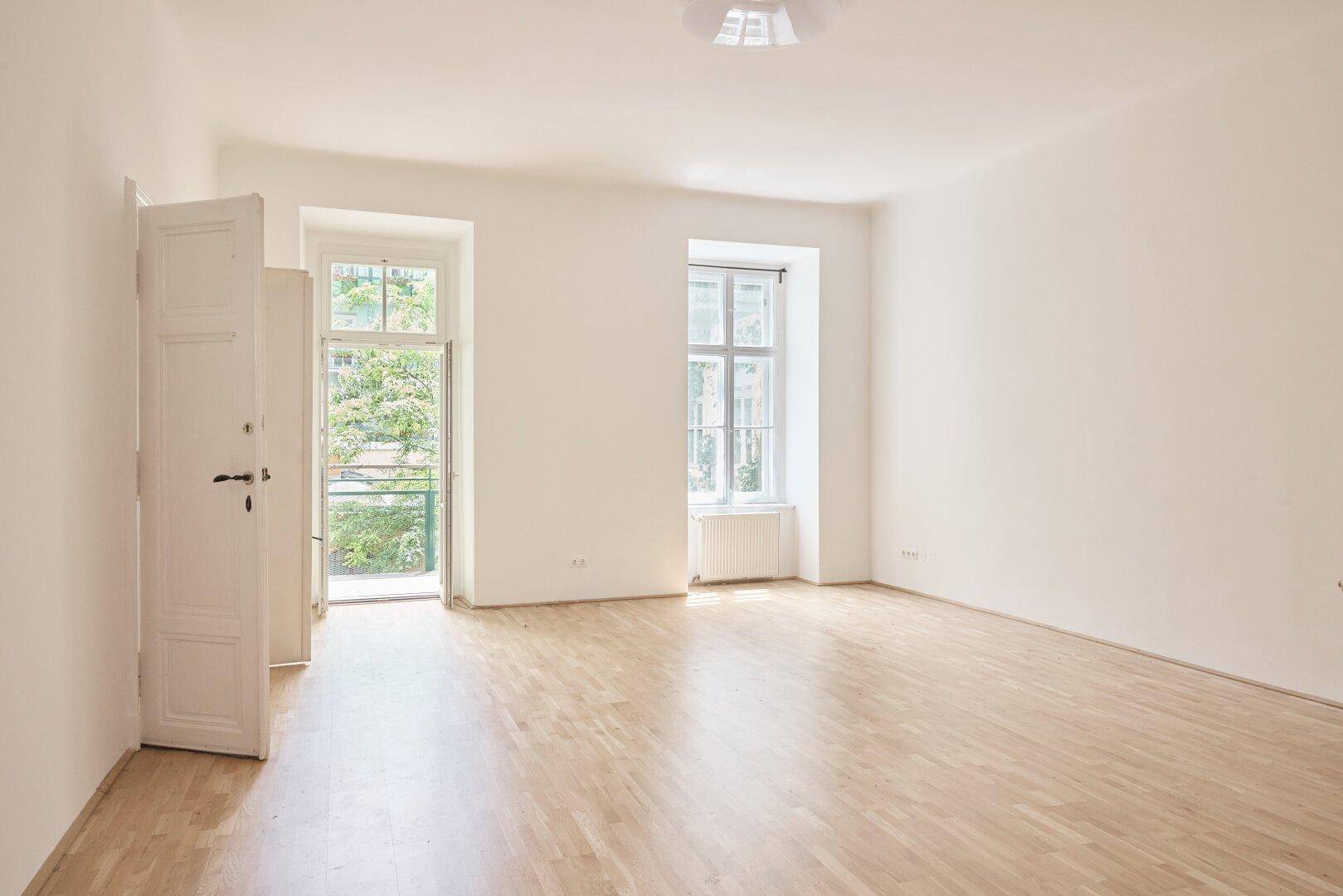 Helles, leeres Zimmer mit Holzdielenboden, Fenstern und Zugang zu einem kleinen Balkon mit Grünblick.