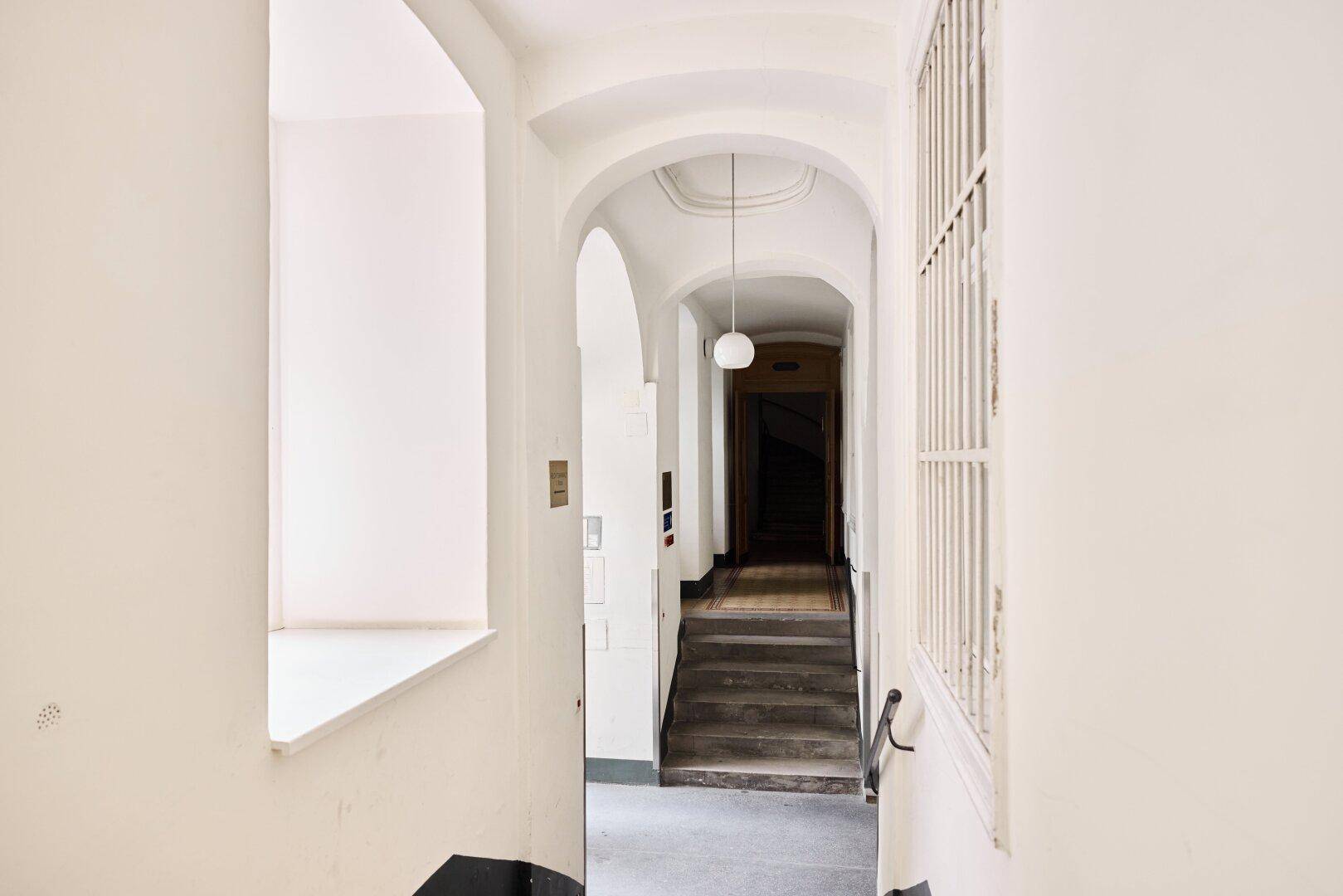 Langer, heller Flur mit Bögen, Treppen und einem Fenster mit Gitter in einem Altbau.