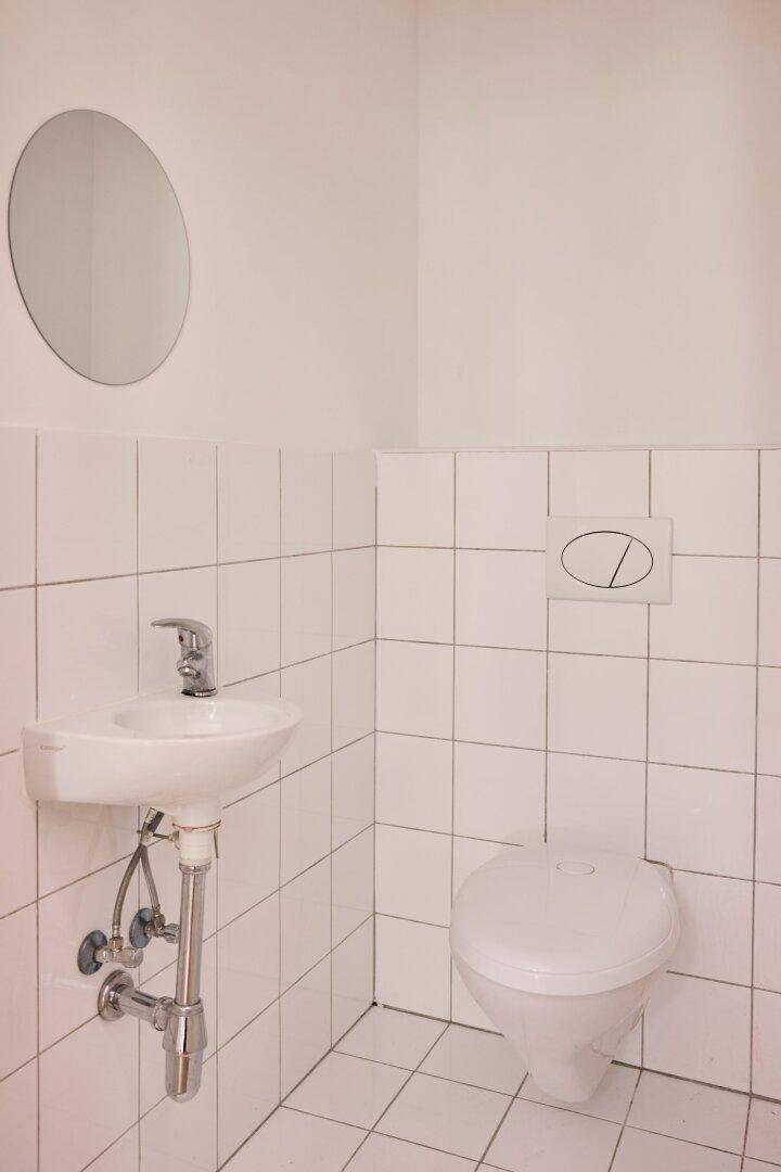 Kleines, hell gefliestes Gäste-WC mit Toilette, Waschbecken und rundem Spiegel.