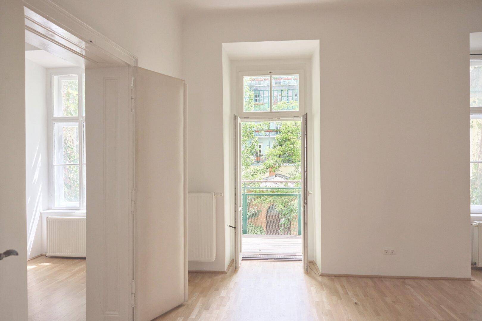 Geräumiges Zimmer mit Holzdielenboden, weiß gestrichenen Wänden und Balkonzugang mit Blick ins Grüne.
