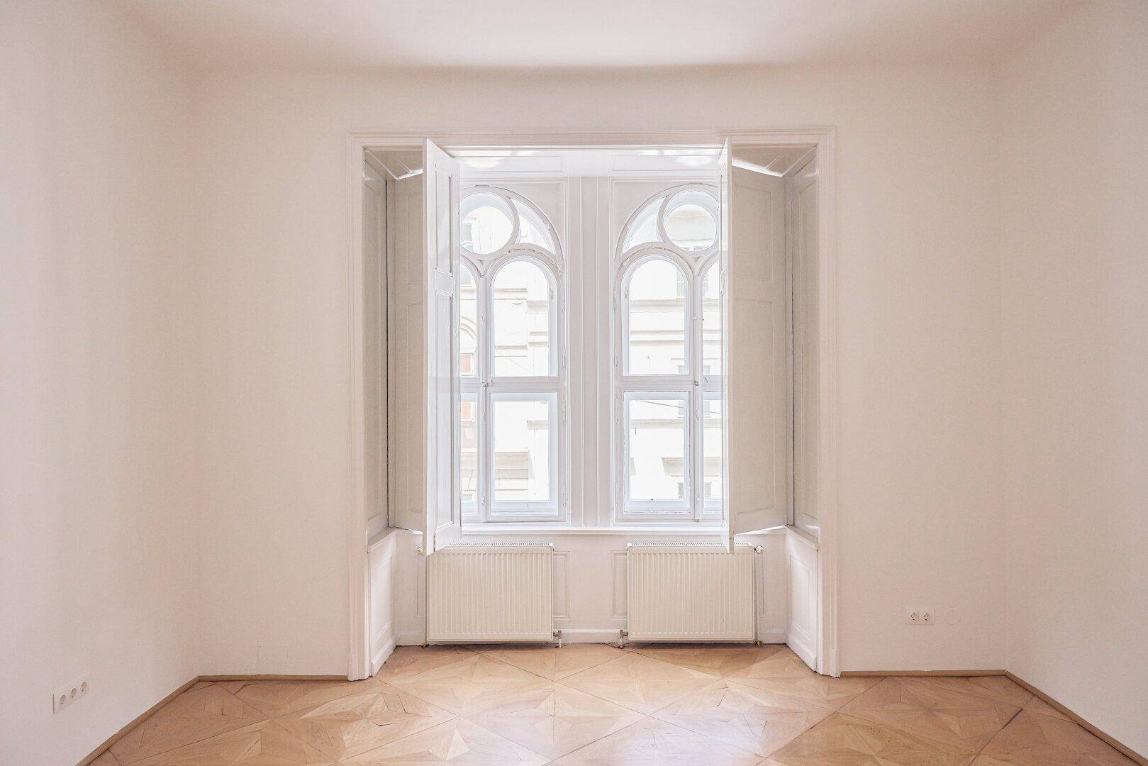 Helles Zimmer mit kunstvollen Bogenfenstern, Parkettboden und weißen Wänden, bereit zur individuellen Gestaltung.