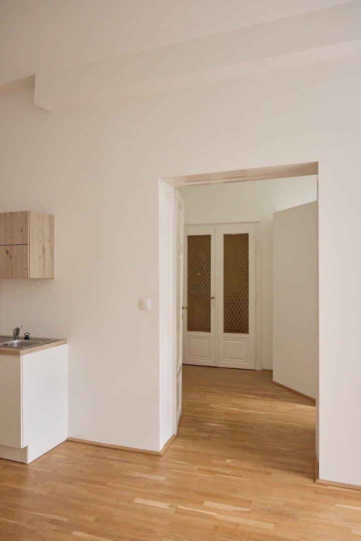 Heller Raum mit Holzdielenboden und einer kleinen Küchenzeile mit Spüle und Oberschrank aus Holz.