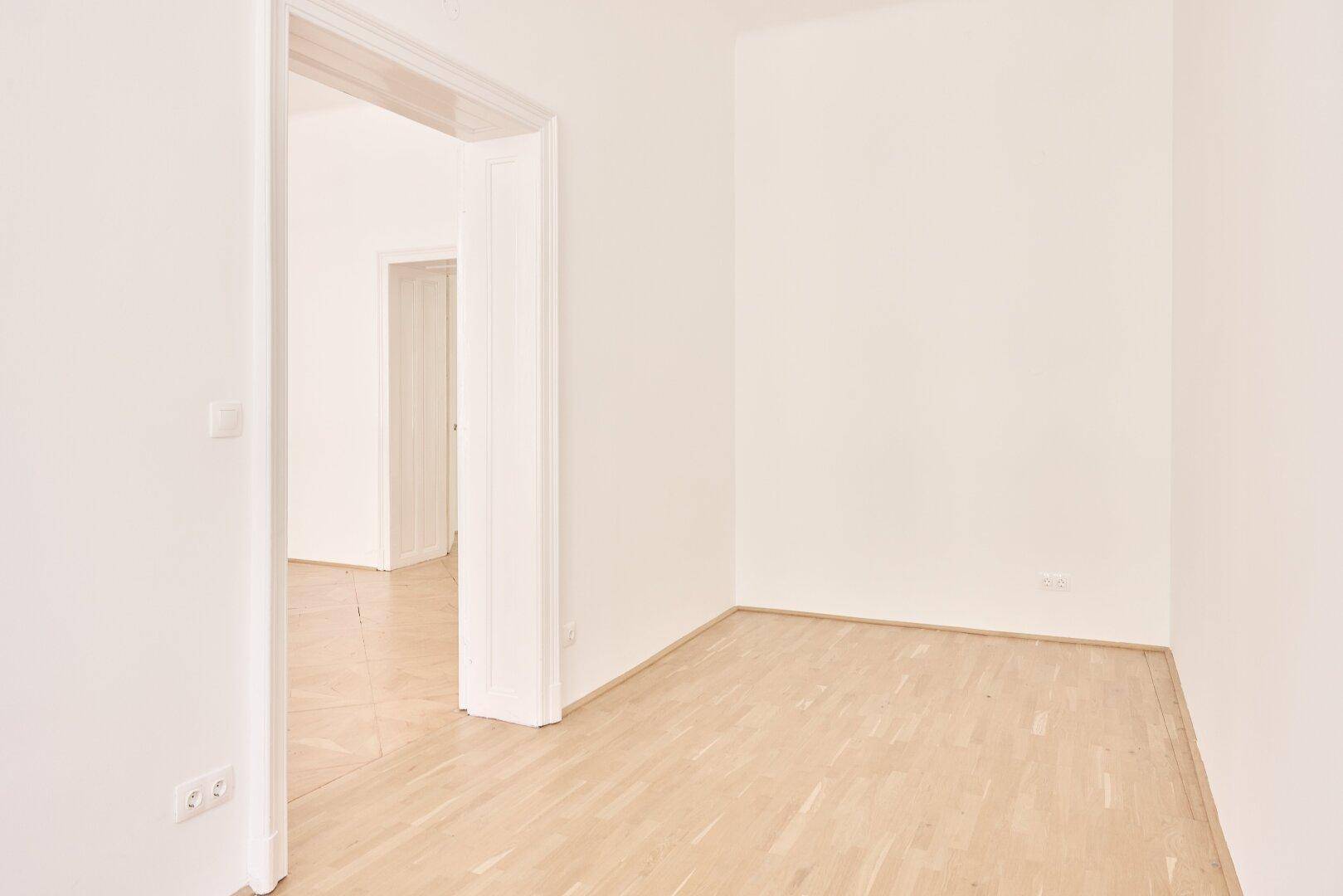 Leeres Zimmer mit hellem Fischgrätparkettboden und einem offenen Durchgang zu einem angrenzenden Raum.