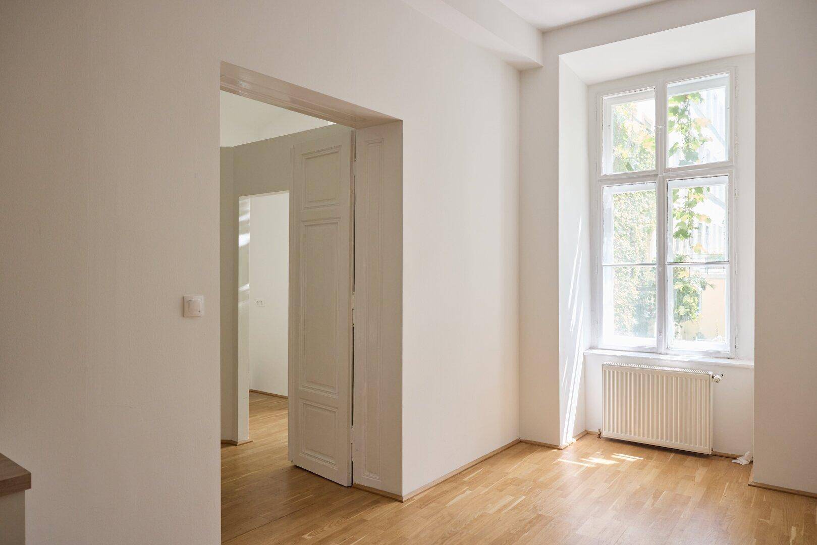 Heller, leerer Raum mit Holzdielenboden, weiß gestrichenen Wänden und einem Fenster mit Heizkörper.