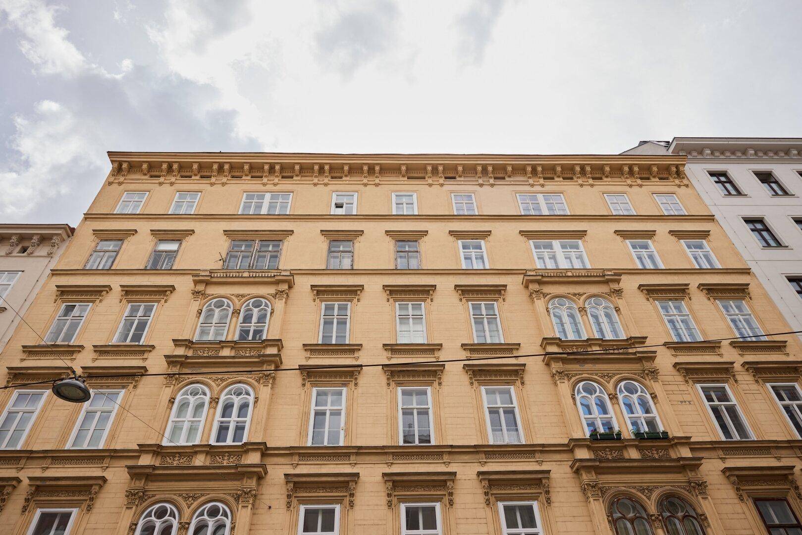 Große historische Gebäudefassade mit vielen Fenstern und dekorativen Elementen unter bewölktem Himmel.
