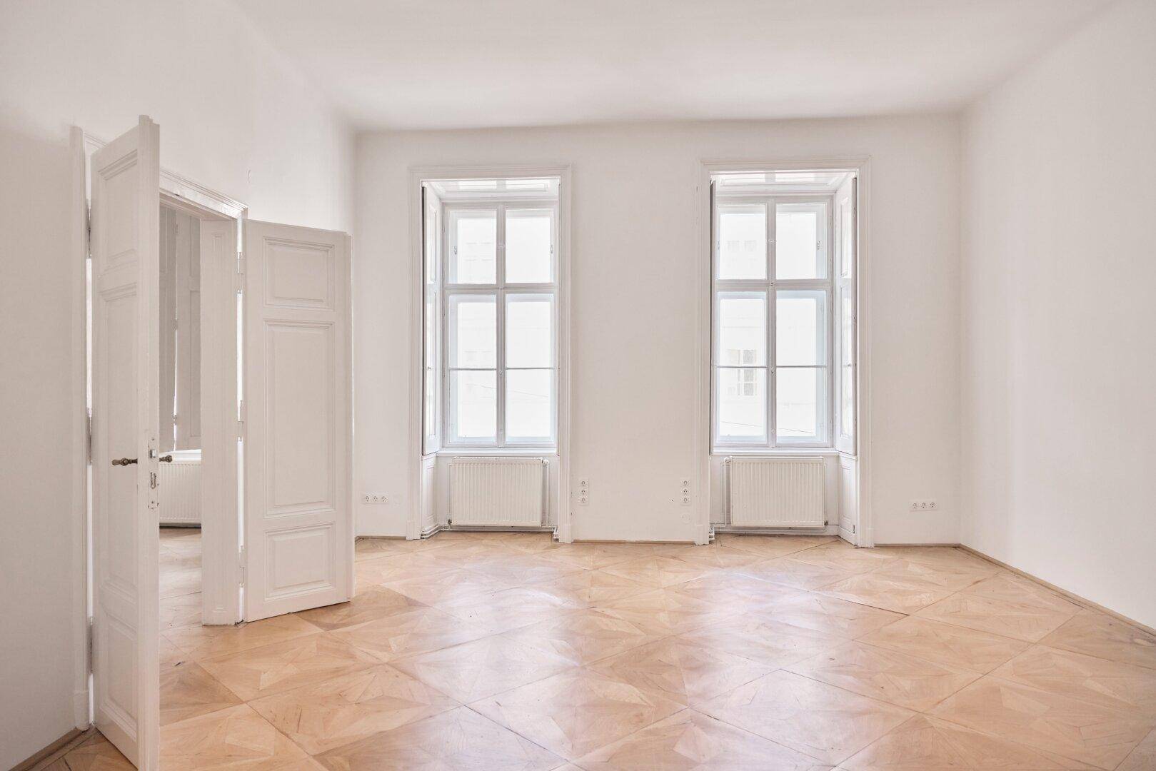Geräumiges, helles Zimmer mit Fischgrätparkettboden, zwei großen Fenstern und offenem Durchgang.