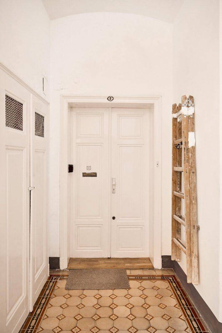 Eingangsbereich eines Gebäudes mit gemusterten Fliesen, weißer Doppeltür und Holzleiter.