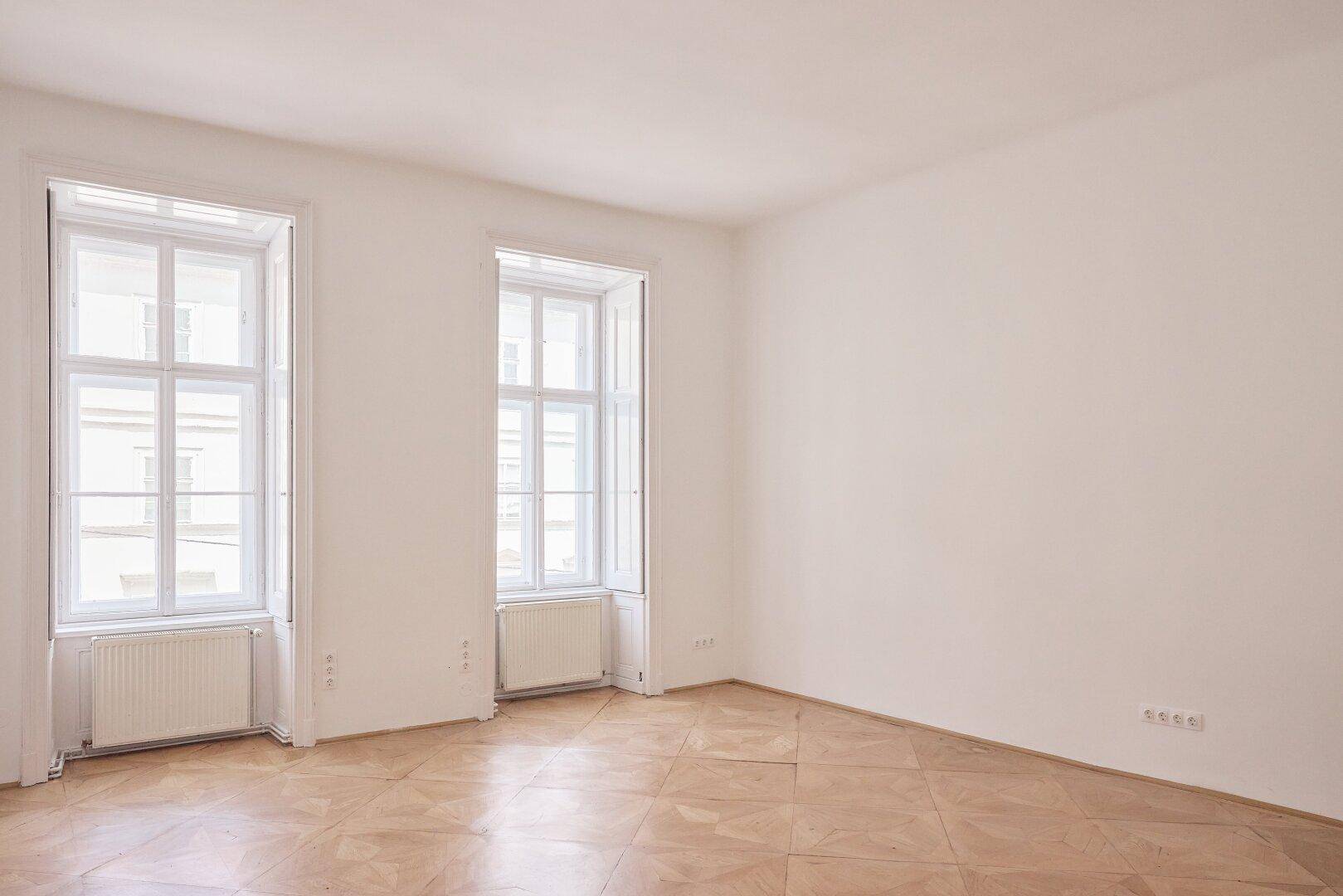 Helles, leeres Zimmer mit Fischgrätparkettboden, weißen Wänden und zwei großen Fenstern.