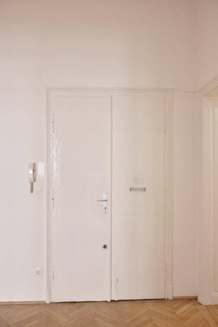 Weiße Eingangstür mit Gegensprechanlage und Briefschlitz in einem hellen Flur mit Parkettboden.