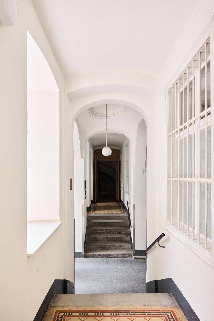 Langer, heller Flur mit Bögen, Treppen und einem Fenster mit Gitter in einem Altbau.