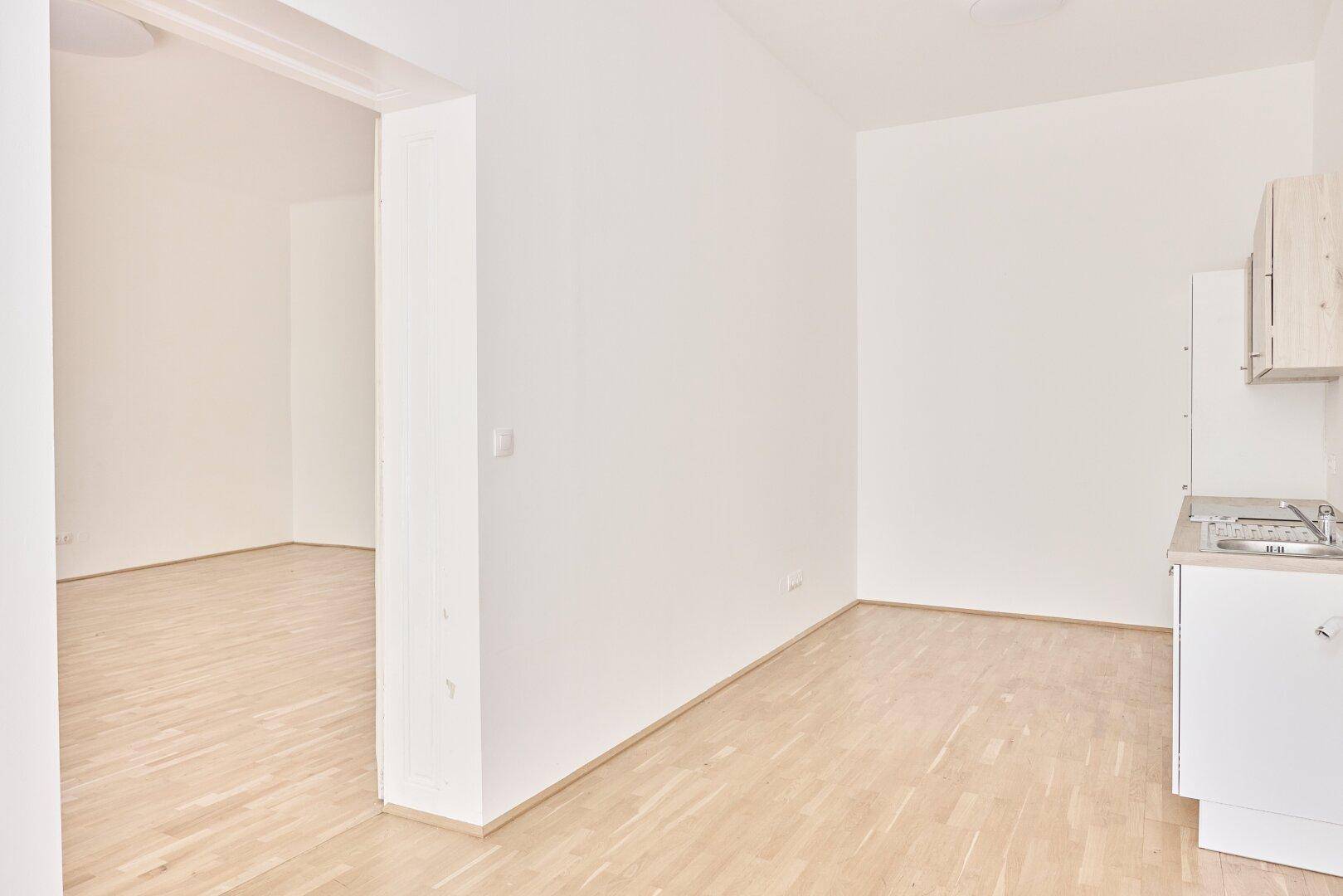 Heller Raum mit Holzdielenboden und einer kleinen Küchenzeile, offen zu einem weiteren Zimmer.