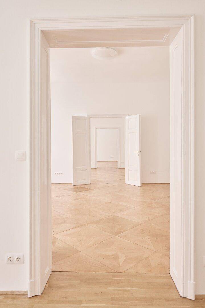 Blick durch eine Reihe offener Türen in helle, leere Räume mit klassischem Parkettboden und hohen Decken.