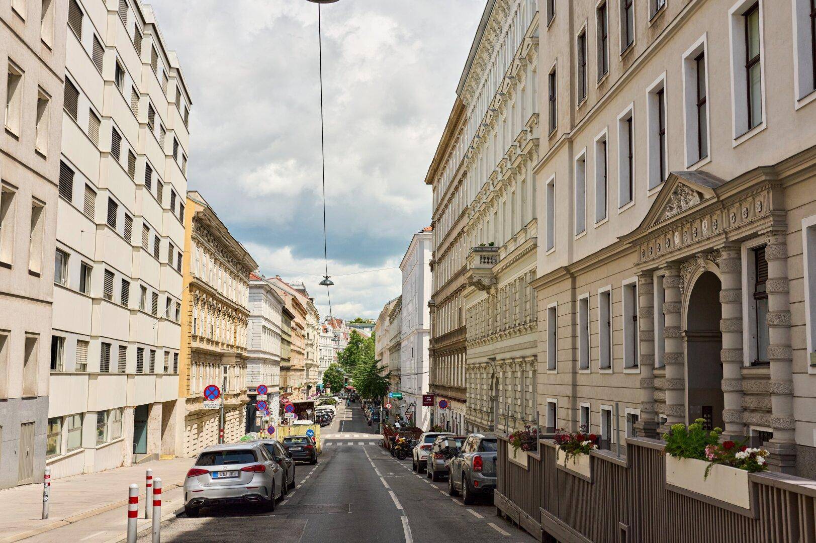 Städtische Straße mit historischen Gebäuden, geparkten Fahrzeugen und bewölktem Himmel.