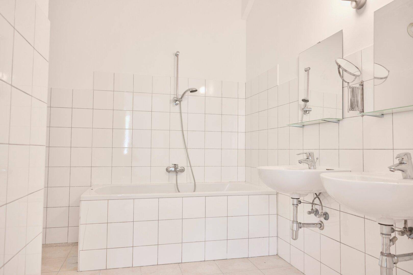 Helles Badezimmer mit weißen Fliesen, Badewanne und zwei Waschbecken mit Spiegeln.