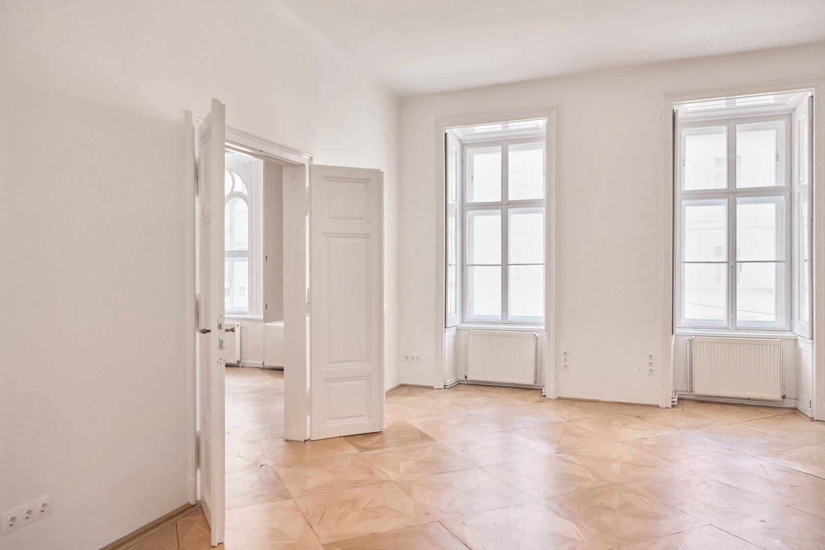 Geräumiger, heller Raum mit hohen Fenstern, Parkettboden und mehreren Türen.