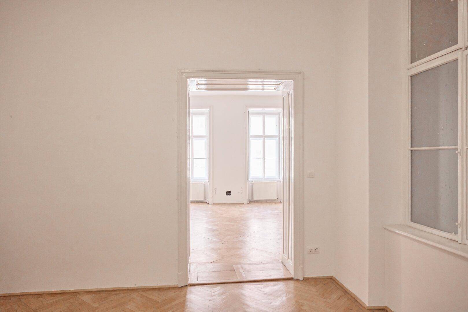 Heller Raum mit offenem Durchgang zu einem weiteren Zimmer mit Fenstern.